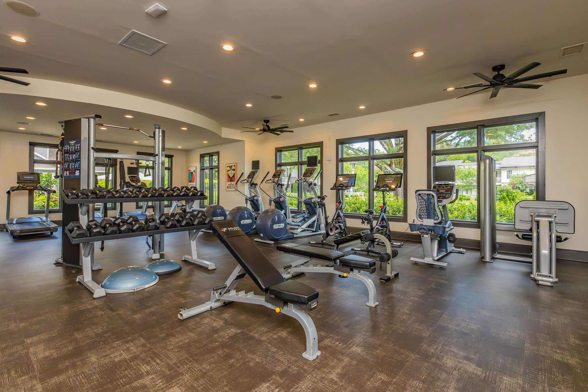A modern gym interior featuring exercise equipment including dumbbells, weight benches, treadmills, and stationary bikes. Brightly lit with large windows showcasing greenery outside, creating a spacious and inviting workout environment.