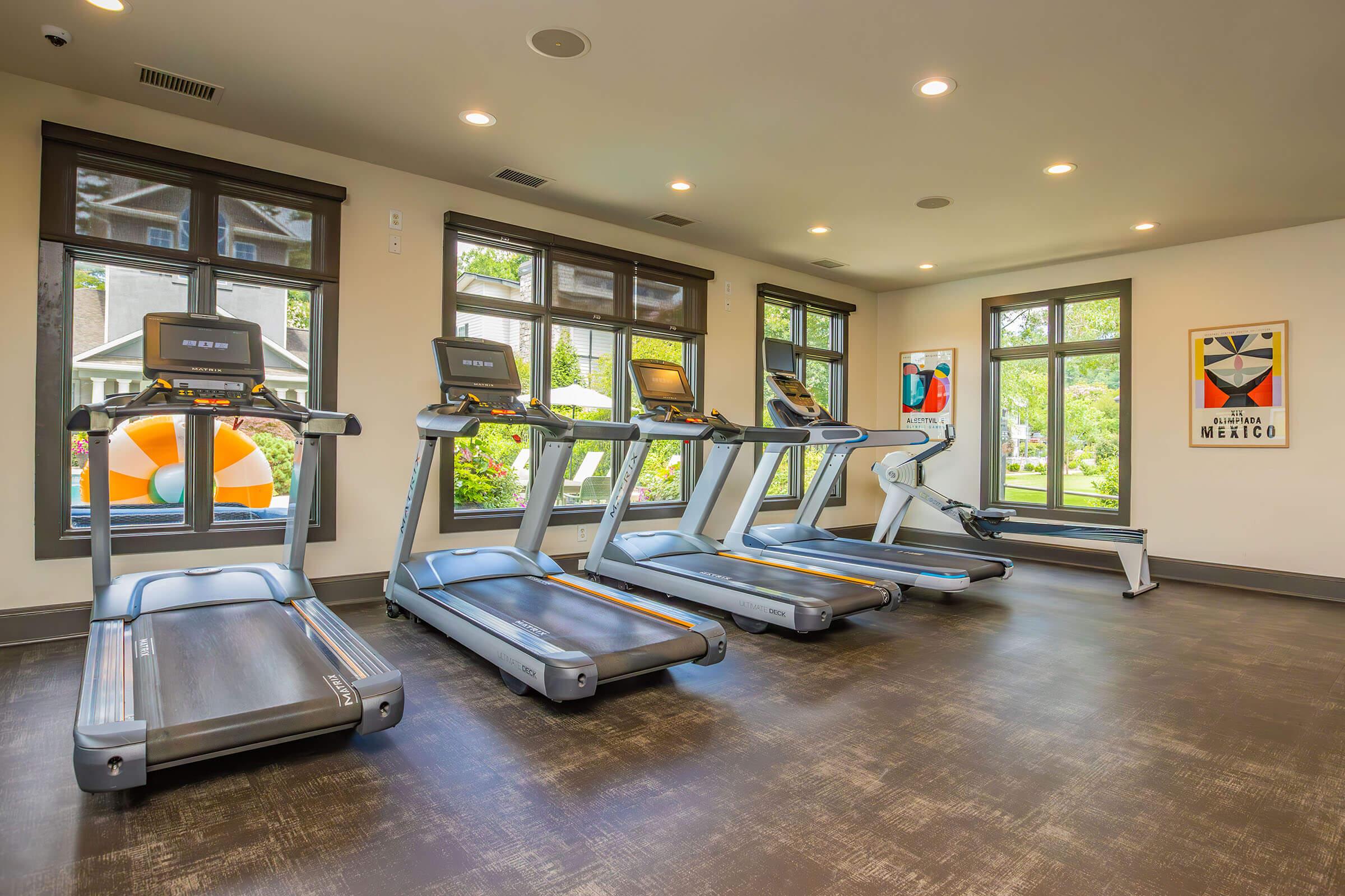 A modern fitness room featuring four treadmills lined up against a wall of large windows, allowing natural light to enter. In the background, there is a colorful beach ball and framed artwork on the walls, creating a welcoming atmosphere for exercise.