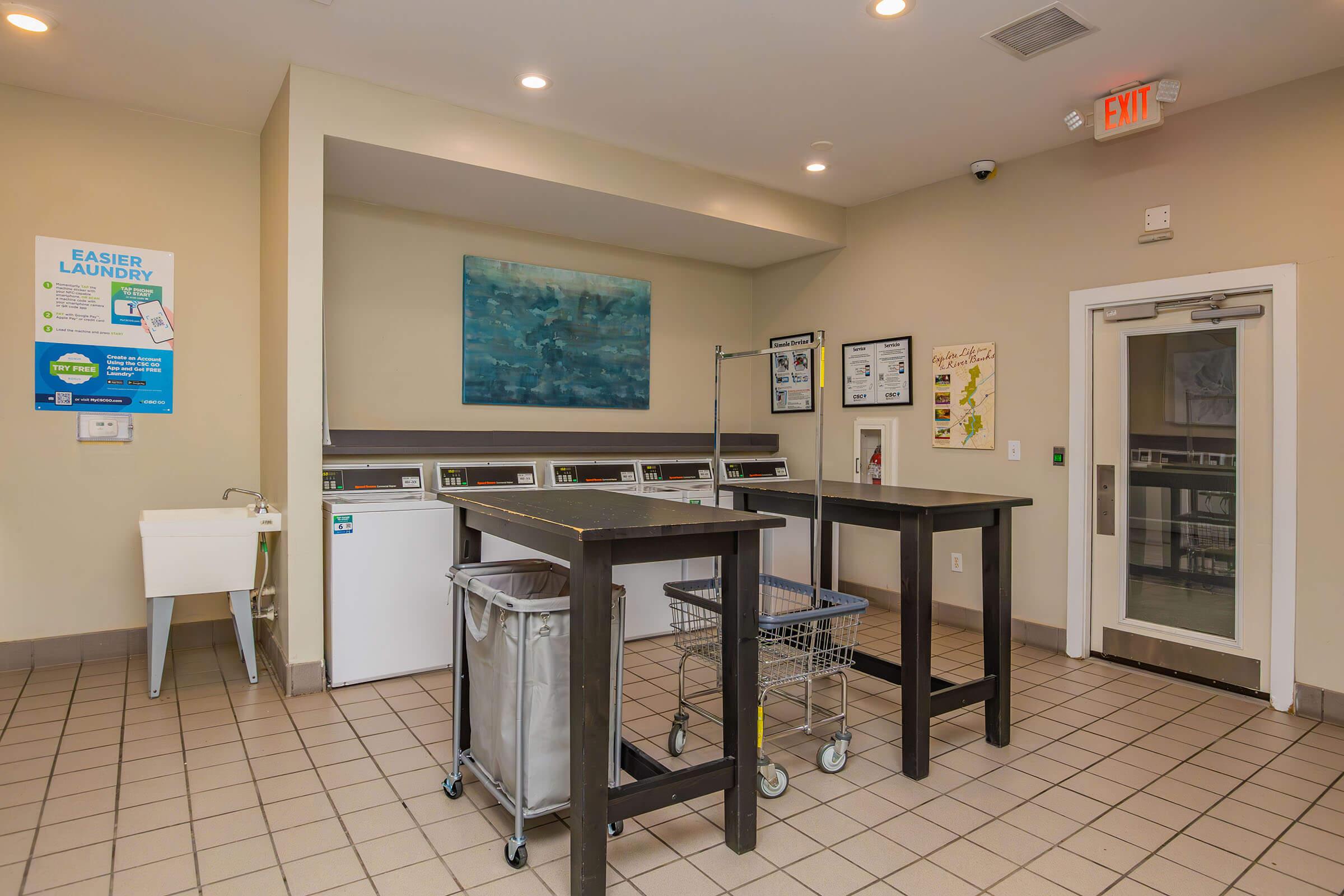 A clean laundry room with several washing machines and drying machines in a well-lit space. There are two tables for folding laundry, a cart for transporting clothes, a utility sink, and informational posters on the walls. The flooring is tiled, and the overall atmosphere is modern and organized.