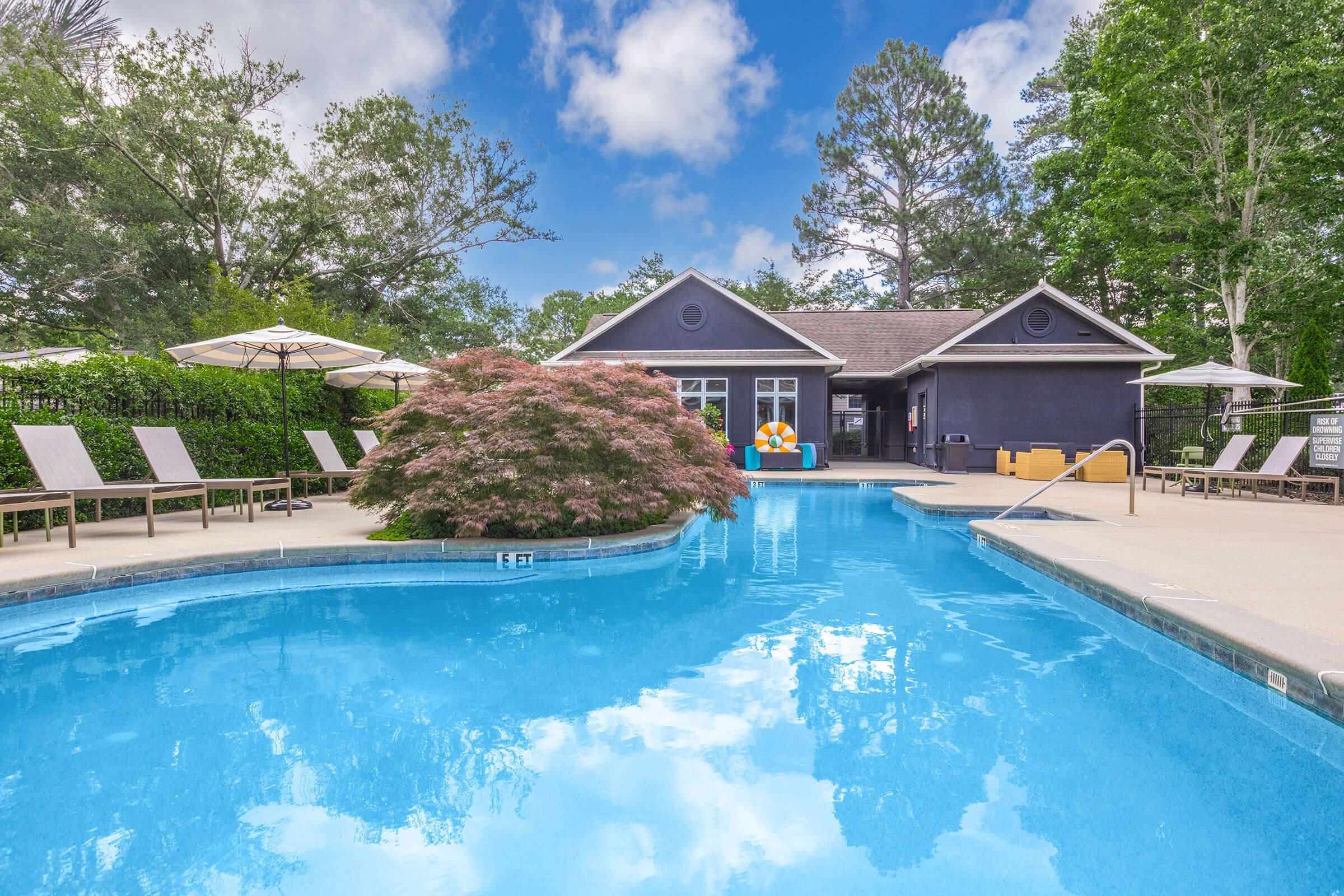 A serene poolside scene featuring a turquoise swimming pool surrounded by lounge chairs and umbrellas. In the background, a modern building with large windows and a colorful pool float. Lush greenery and trees frame the area, creating a relaxing outdoor atmosphere under a partly cloudy sky.