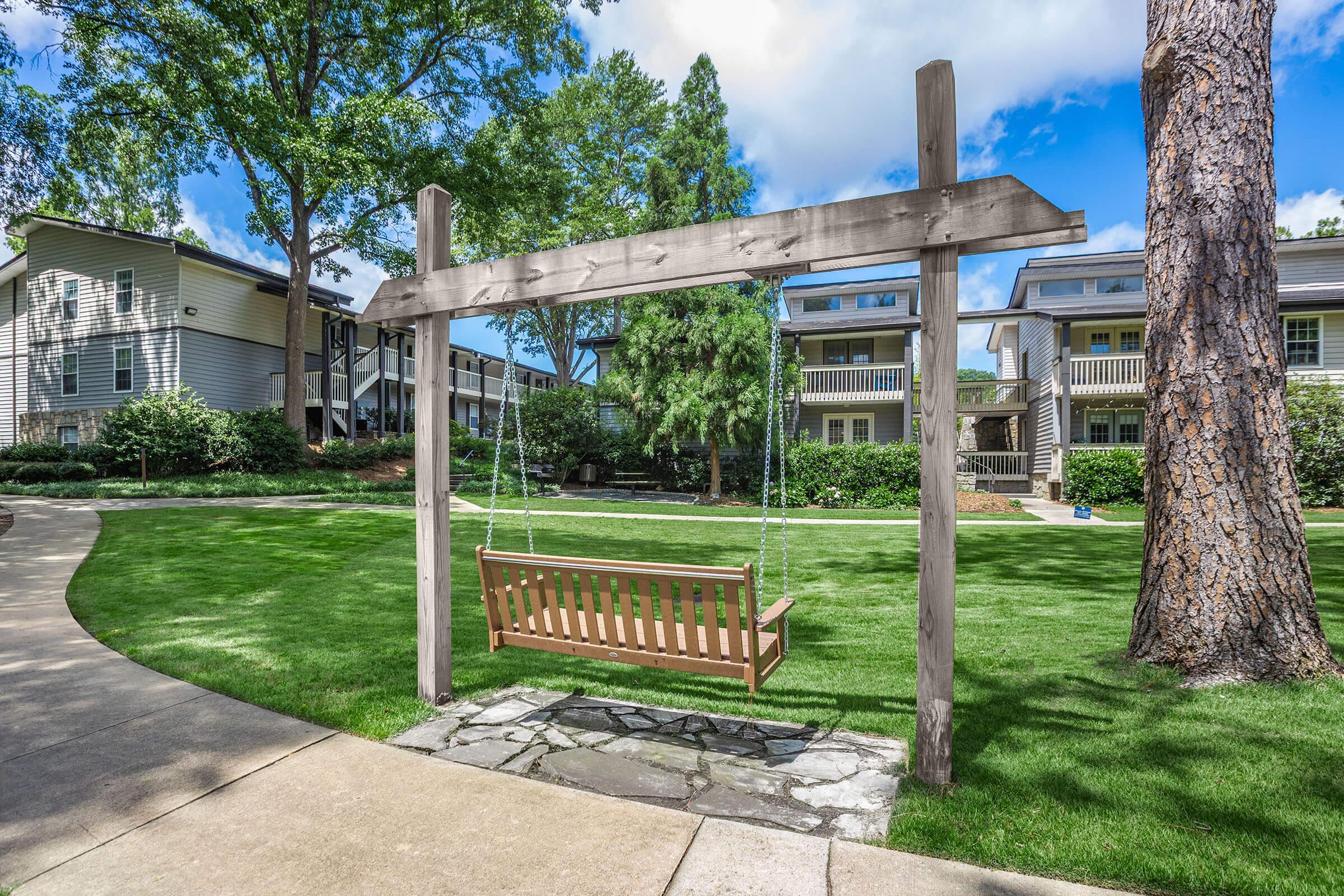 A wooden swing hangs from a sturdy frame in a well-manicured green space. The swing is positioned on a stone base, surrounded by grass and trees. In the background, there are modern apartment buildings and a clear blue sky with wispy clouds. Pathways wind through the landscape.