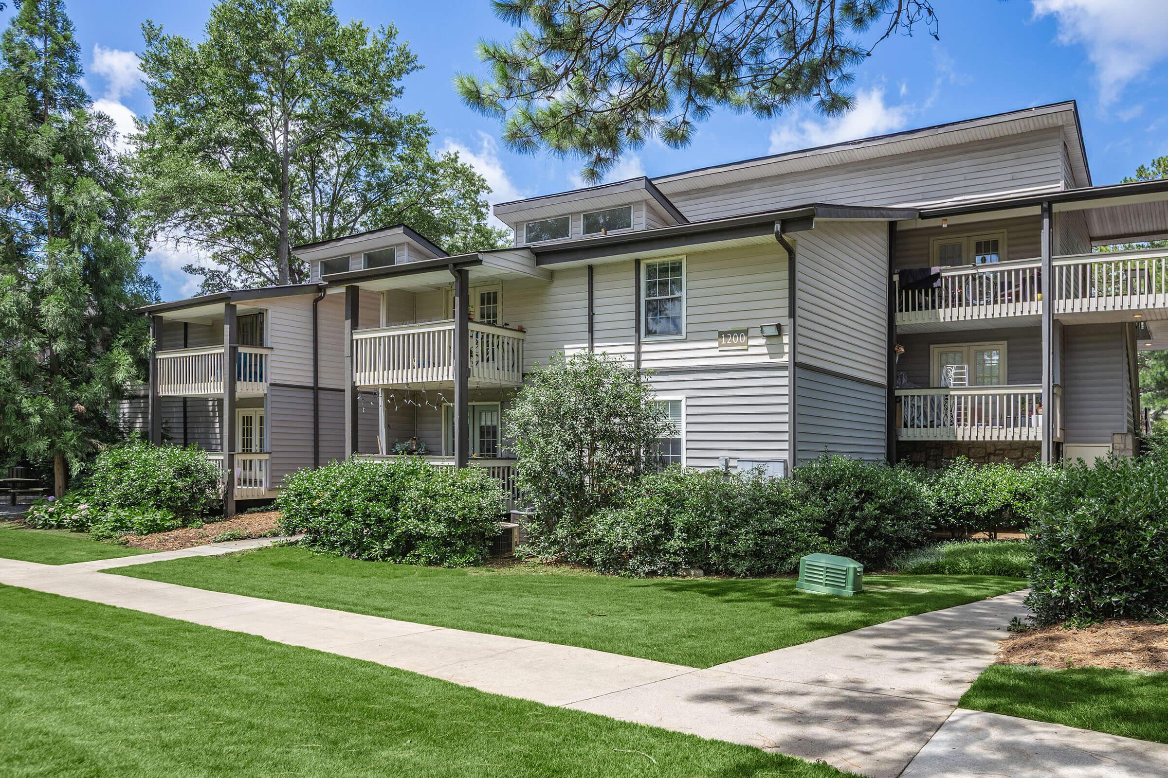 Two-story apartment building with multiple balconies, surrounded by greenery and well-maintained lawns. Pathways lead through the landscaped area, with trees in the background and a clear blue sky overhead.
