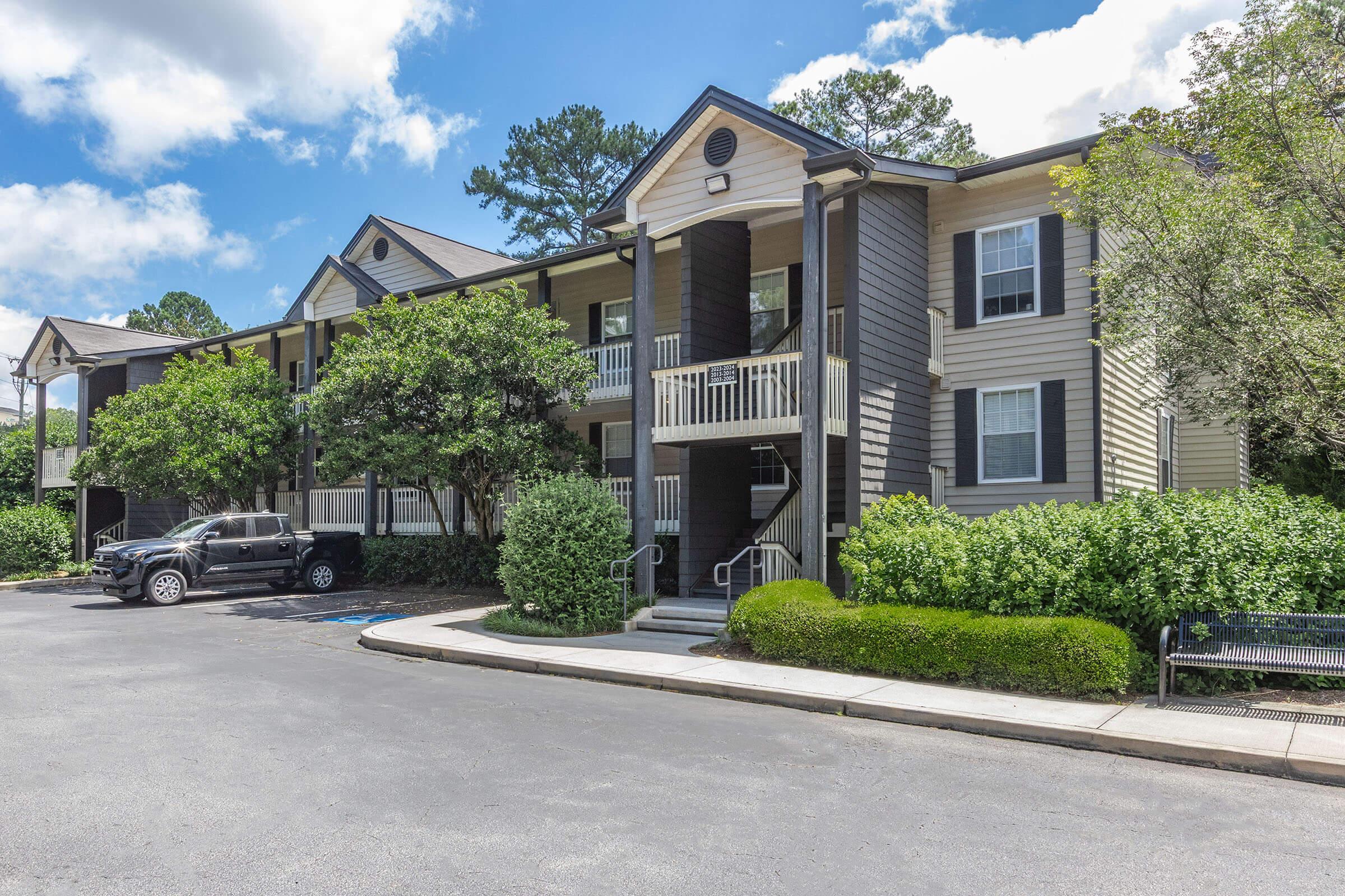 A two-story apartment building featuring a mix of beige and dark brown siding. The facade includes balconies with white railings. Lush green bushes and trees surround the building. A black SUV is parked nearby, and there is a bench in front of the building under a partly cloudy sky.