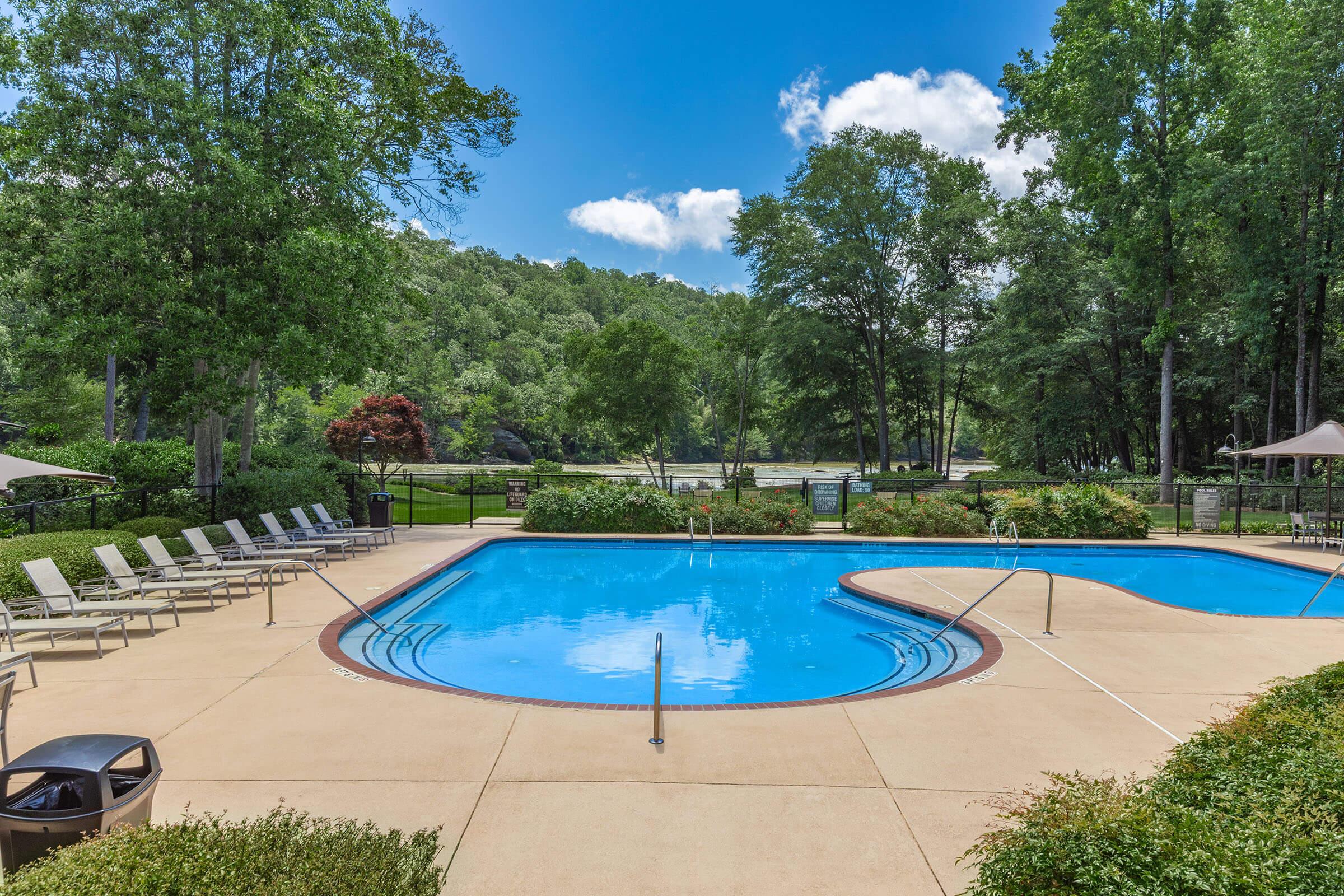A clear blue swimming pool surrounded by lounge chairs, set in a landscaped area with trees and green hills in the background under a bright blue sky with fluffy white clouds.