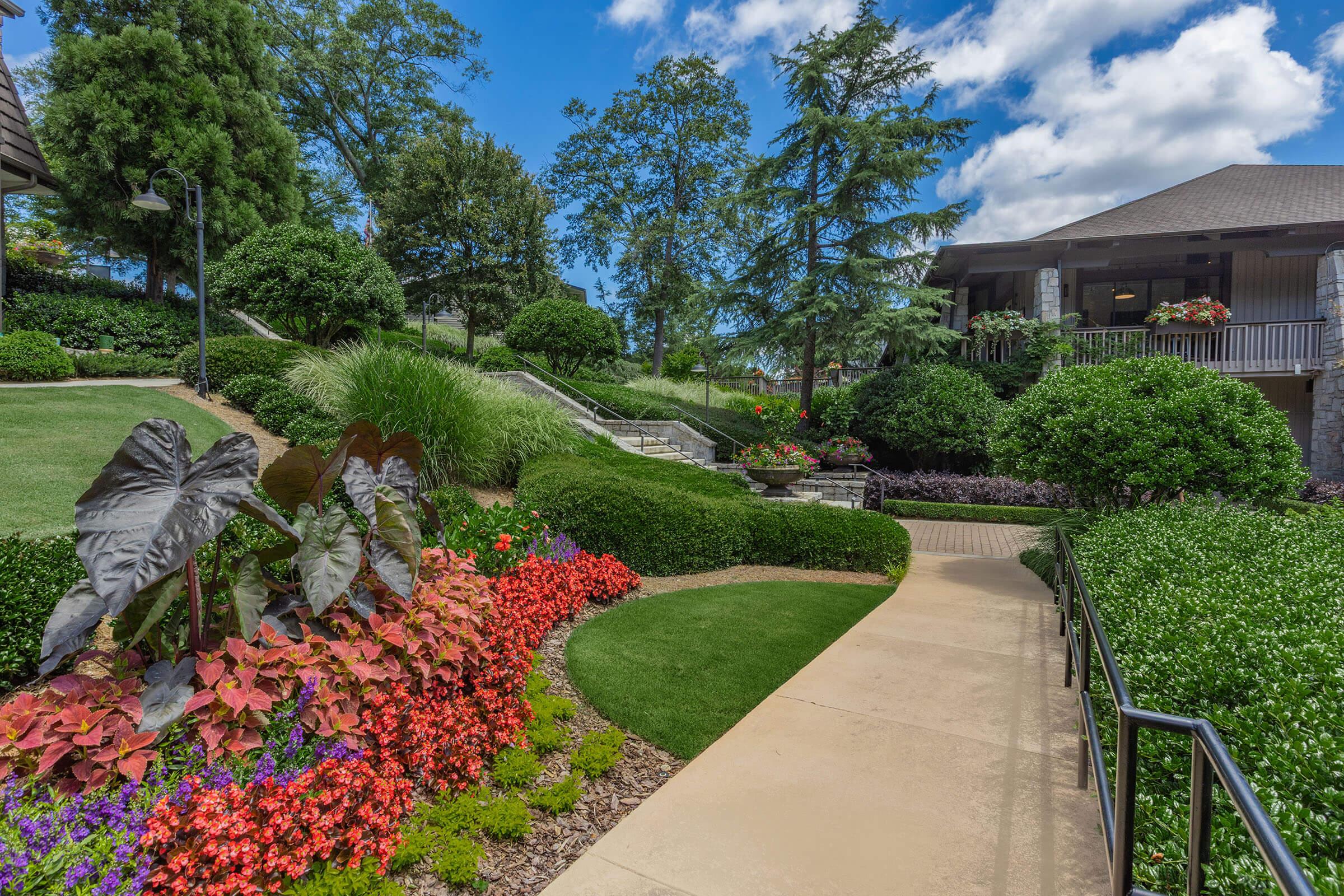 A landscaped pathway lined with vibrant flowers in red, pink, and yellow, leading through lush green shrubs and trees. In the background, a stone building with a porch is visible under a bright blue sky with fluffy white clouds. The scene conveys a serene and inviting outdoor space.