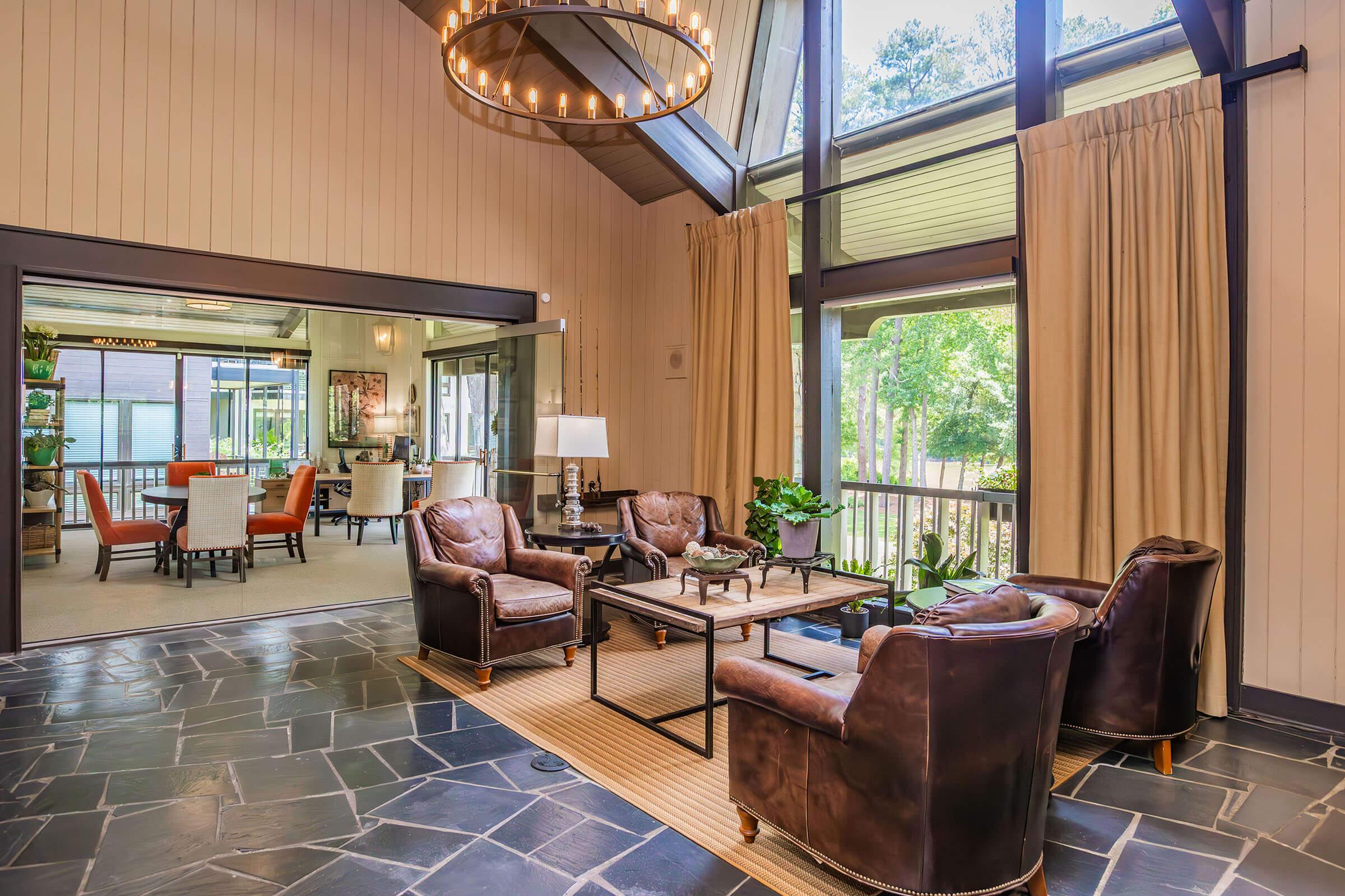 A cozy interior featuring a well-lit sitting area with leather armchairs, a coffee table, and a modern chandelier. Large windows allow natural light, showcasing outdoor greenery. Nearby, a glimpse of another room with elegant seating and decor can be seen through an open doorway.