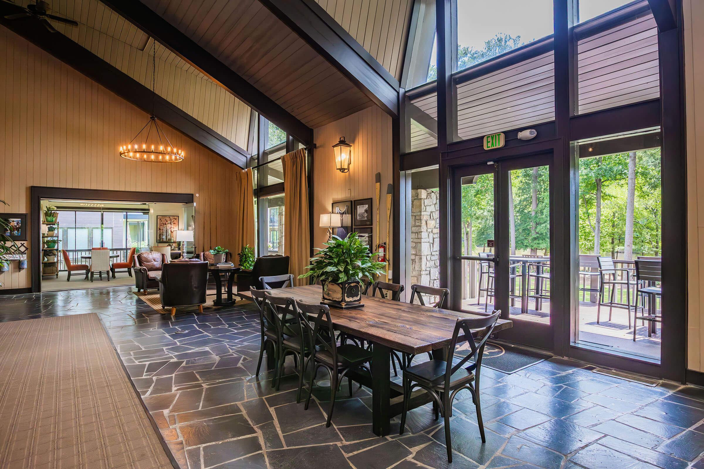 A spacious indoor area featuring a large wooden dining table surrounded by black chairs, with a stone floor and tall, vaulted ceilings. Large windows let in natural light, offering a view of greenery outside. The space includes a seating area with comfortable couches and decorative lighting.