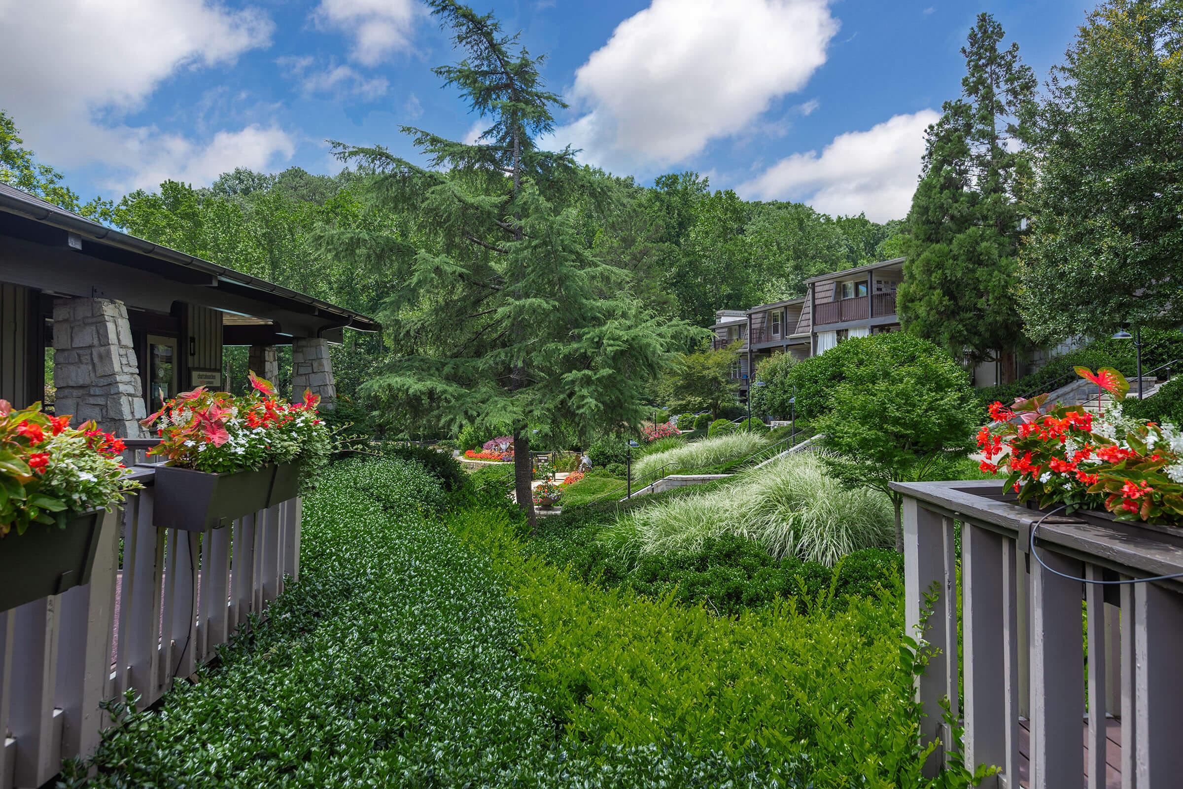 Lush garden scene featuring vibrant flower pots along a pathway, leading to a well-maintained landscape with trees and green foliage. In the background, modern buildings are partially visible under a bright blue sky with fluffy white clouds, creating a serene and inviting atmosphere.
