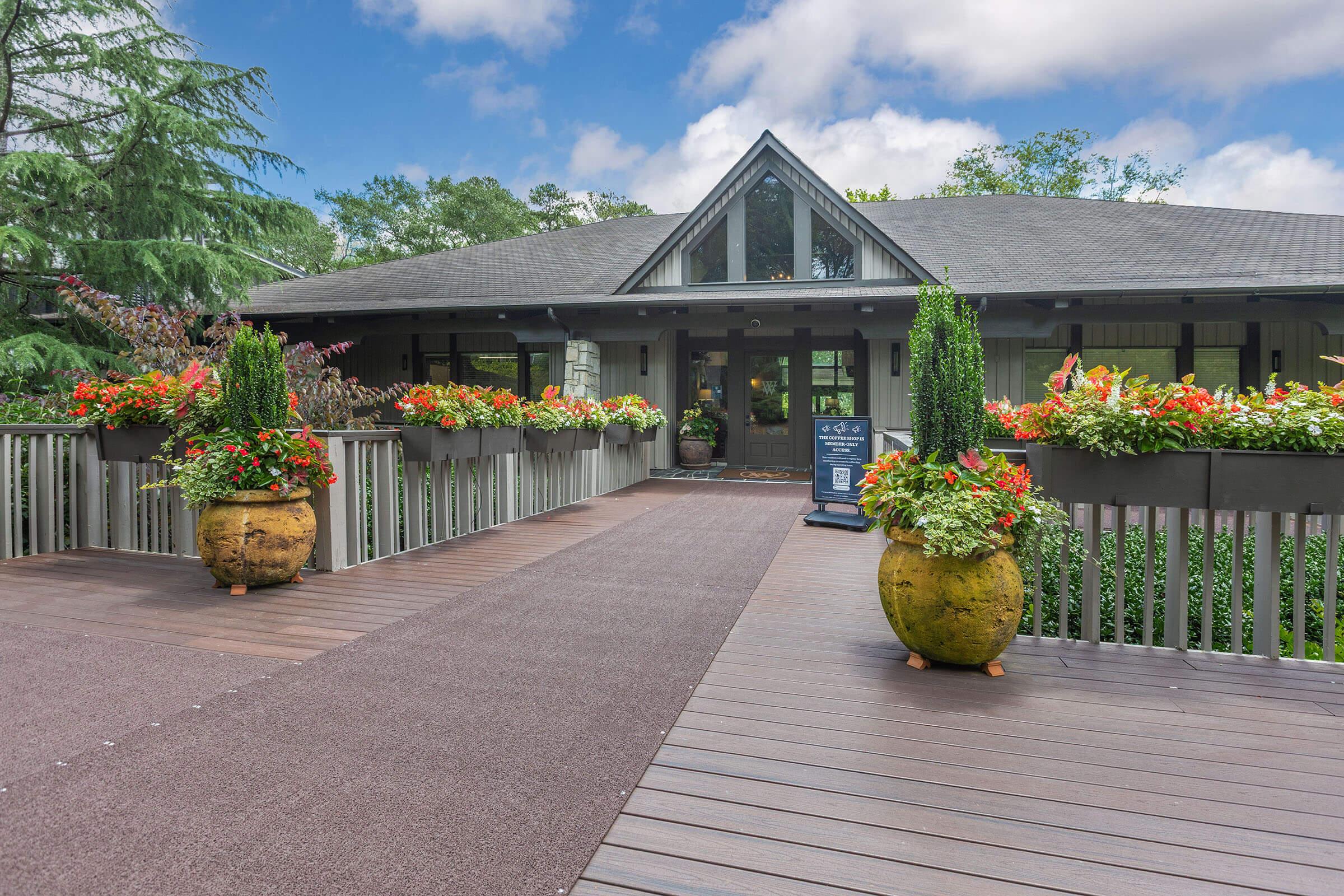 A welcoming entrance to a building with a wooden deck, adorned with vibrant flower pots on either side. The setting features lush greenery in the background and a cloudy blue sky above, creating a picturesque and inviting atmosphere.