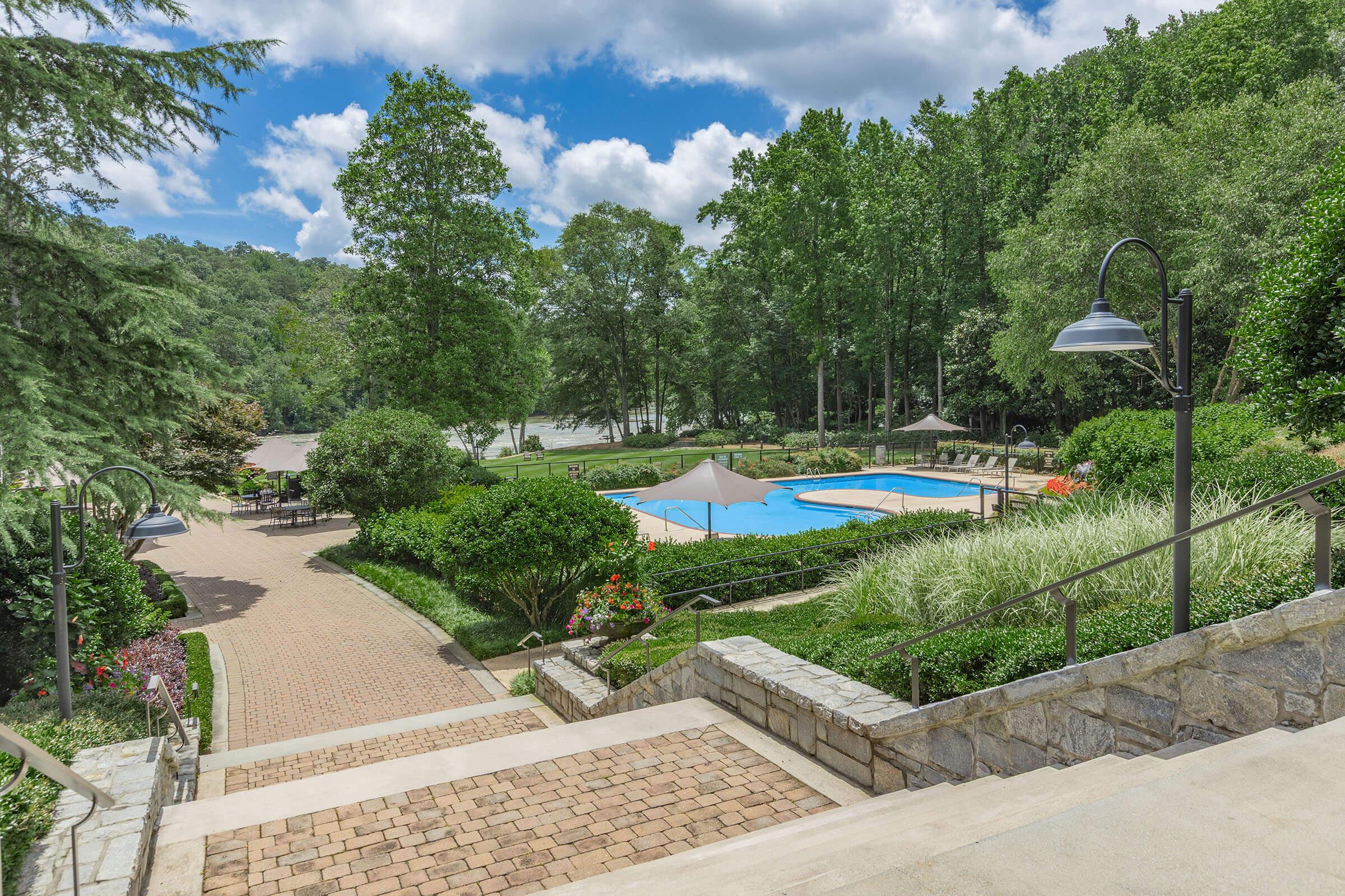 Scenic view of a landscaped resort area featuring a swimming pool surrounded by greenery, lounge chairs, and shaded seating. Stone pathways lead down to the pool area, with vibrant flowers and trees providing a tranquil atmosphere under a partly cloudy sky.