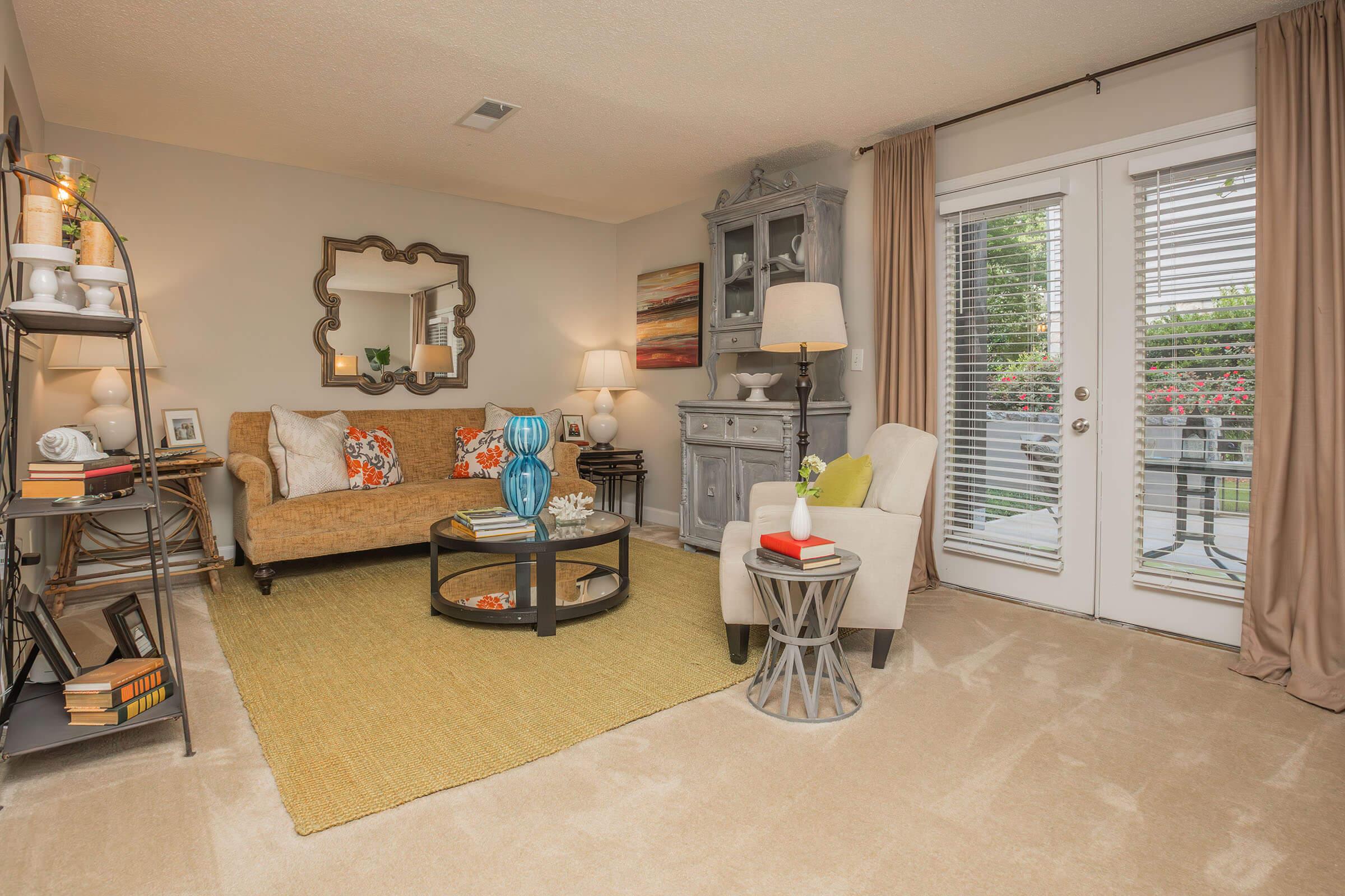 A cozy living room featuring a tan sofa with colorful throw pillows, a decorative mirror on the wall, a glass coffee table, and a light-colored armchair. Natural light streams through sliding glass doors with curtains, and a stylish cabinet is visible in the background, complemented by a textured area rug.