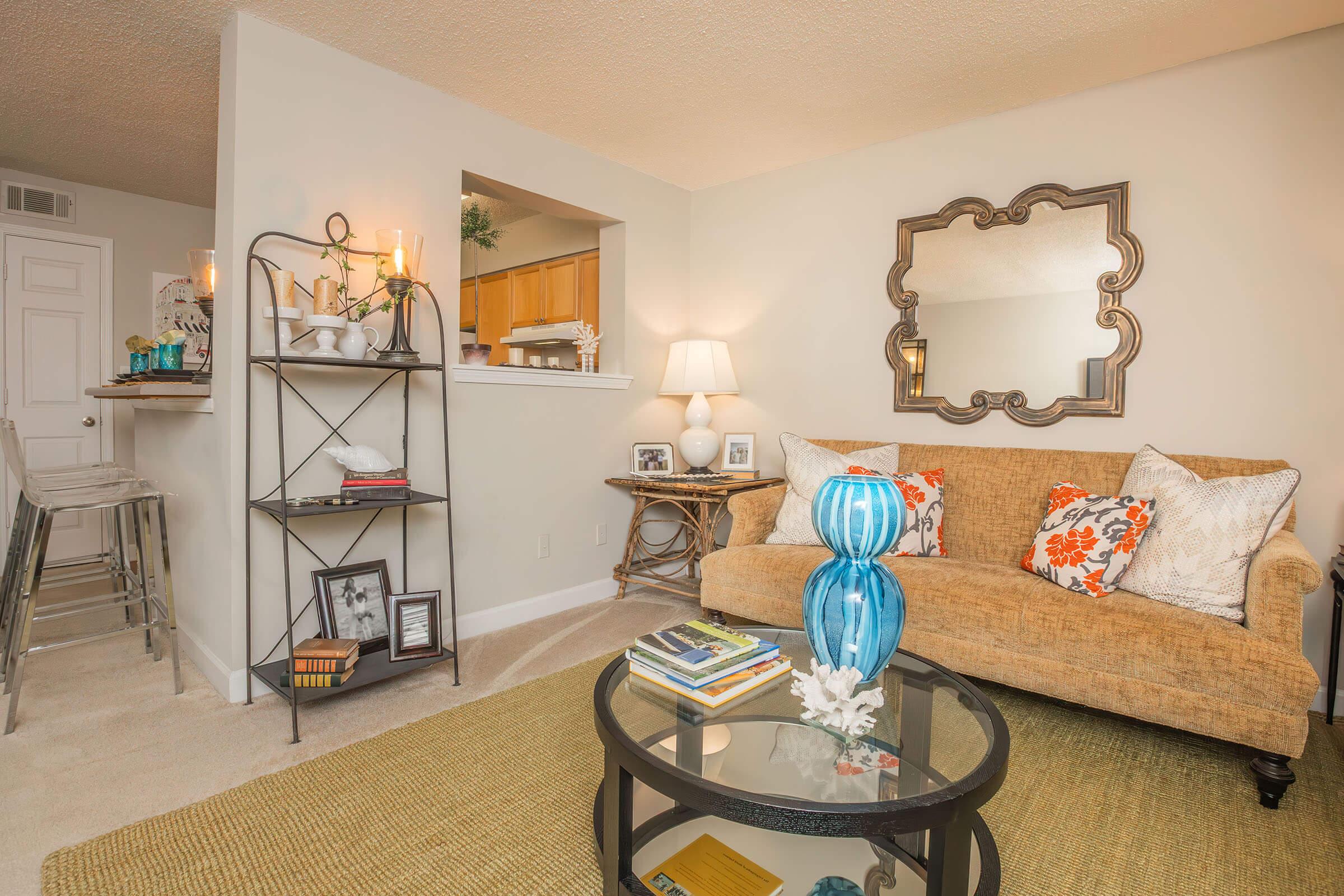A cozy living room featuring a tan sofa with colorful throw pillows, a glass coffee table adorned with decor, and a stylish shelving unit holding books and candles. The walls are light-colored, with a large mirror reflecting the space, and a soft rug adds warmth to the floor. A glimpse of the kitchen is visible in the background.