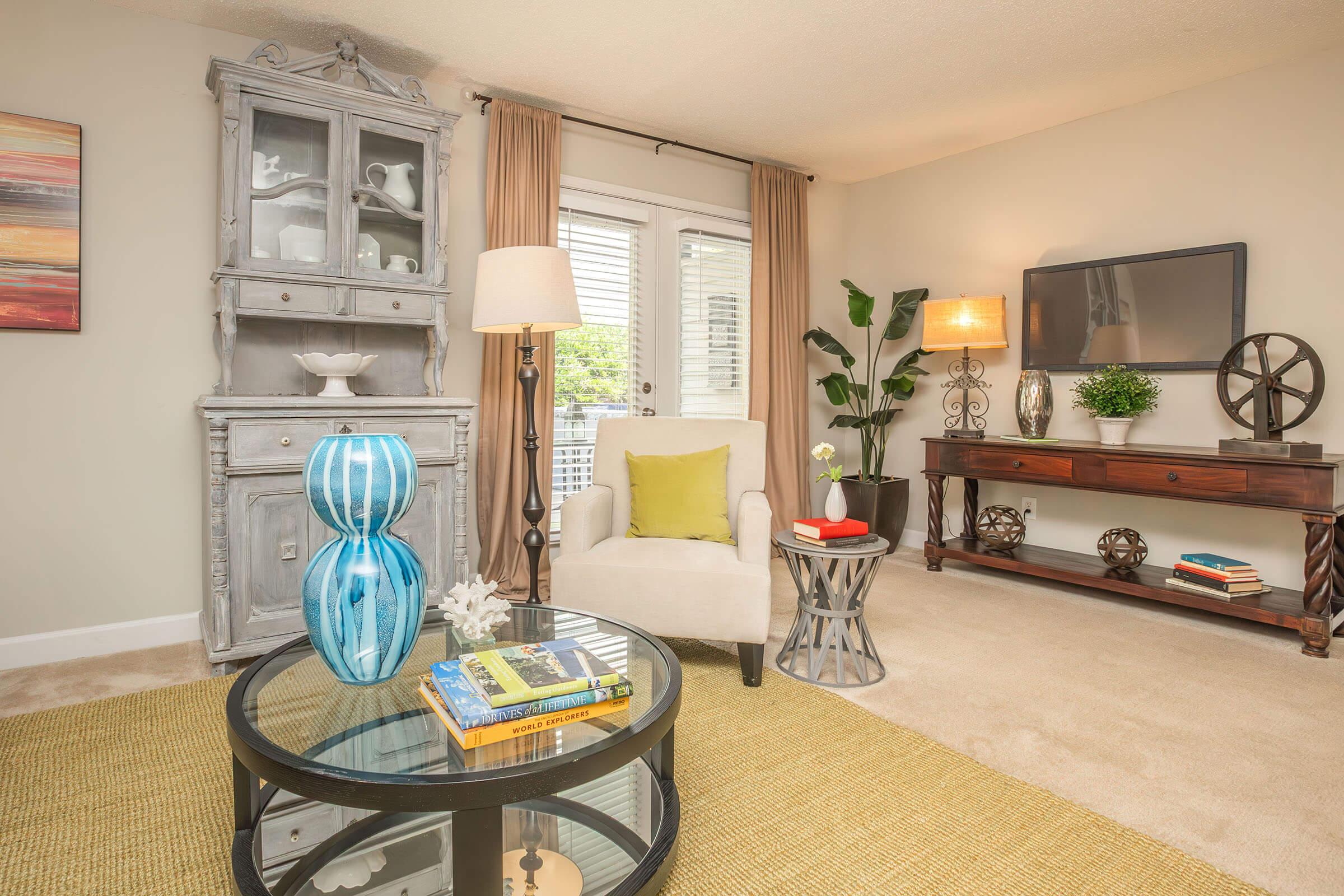A cozy living room featuring a light gray armchair with a yellow throw pillow, a stylish circular coffee table with books and decorative items, a wooden console table with a lamp and plant, and a vintage cabinet. Natural light comes through the curtains, enhancing the inviting atmosphere.