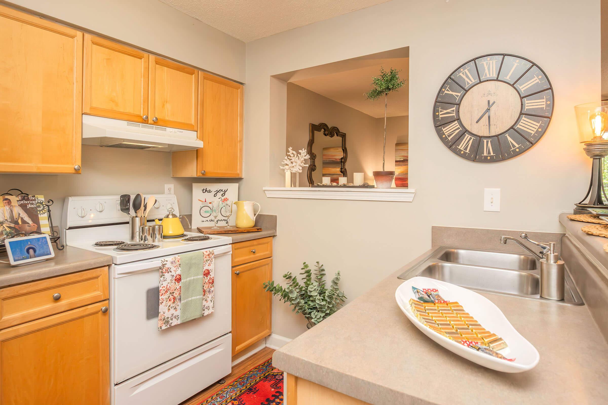 A cozy kitchen featuring wooden cabinets, a white stove, and a sink with modern fixtures. The countertop is adorned with a decorative dish and a kettle. A large clock hangs on the wall, and there’s a small window with plants. The overall ambiance is warm and inviting.
