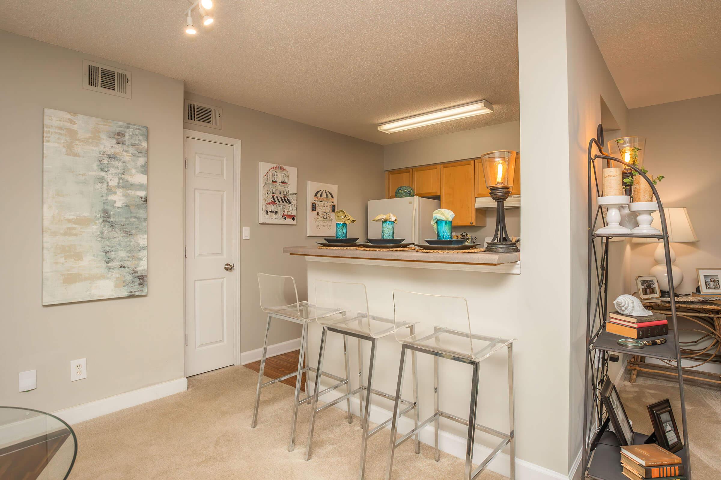 A cozy, modern apartment interior featuring a small kitchen bar with three clear stools. Soft beige walls complement light cabinetry and decorative elements. A wall art piece adds color, and a shelf displays books and decor. Warm lighting creates an inviting atmosphere.