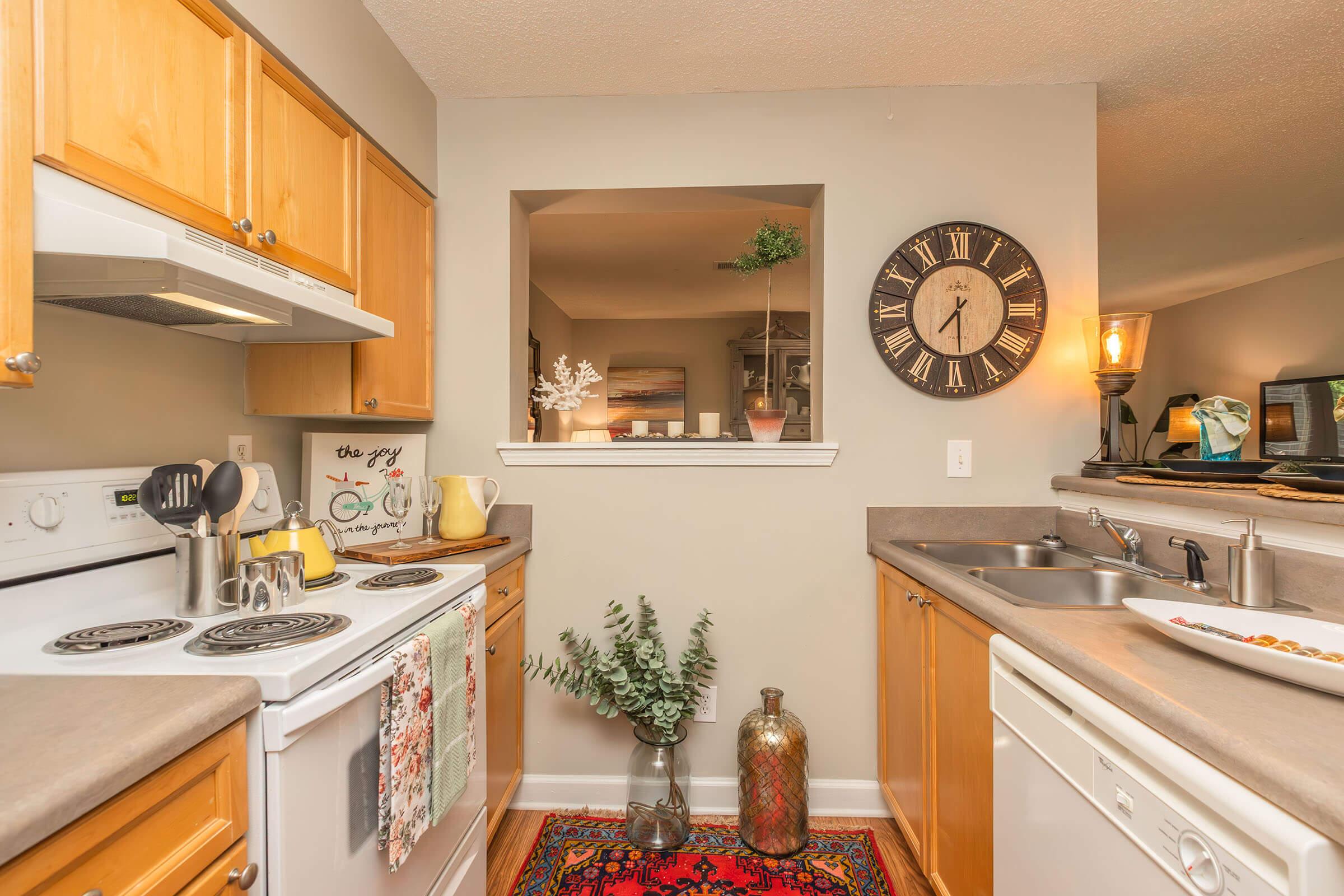 A cozy kitchen featuring wooden cabinets, a white stove and dishwasher. A large clock is mounted on the wall, with a small window opening to a dining area. Decorative plants and a colorful rug add warmth to the space. The countertop is arranged with kitchen essentials and a few decorative items.