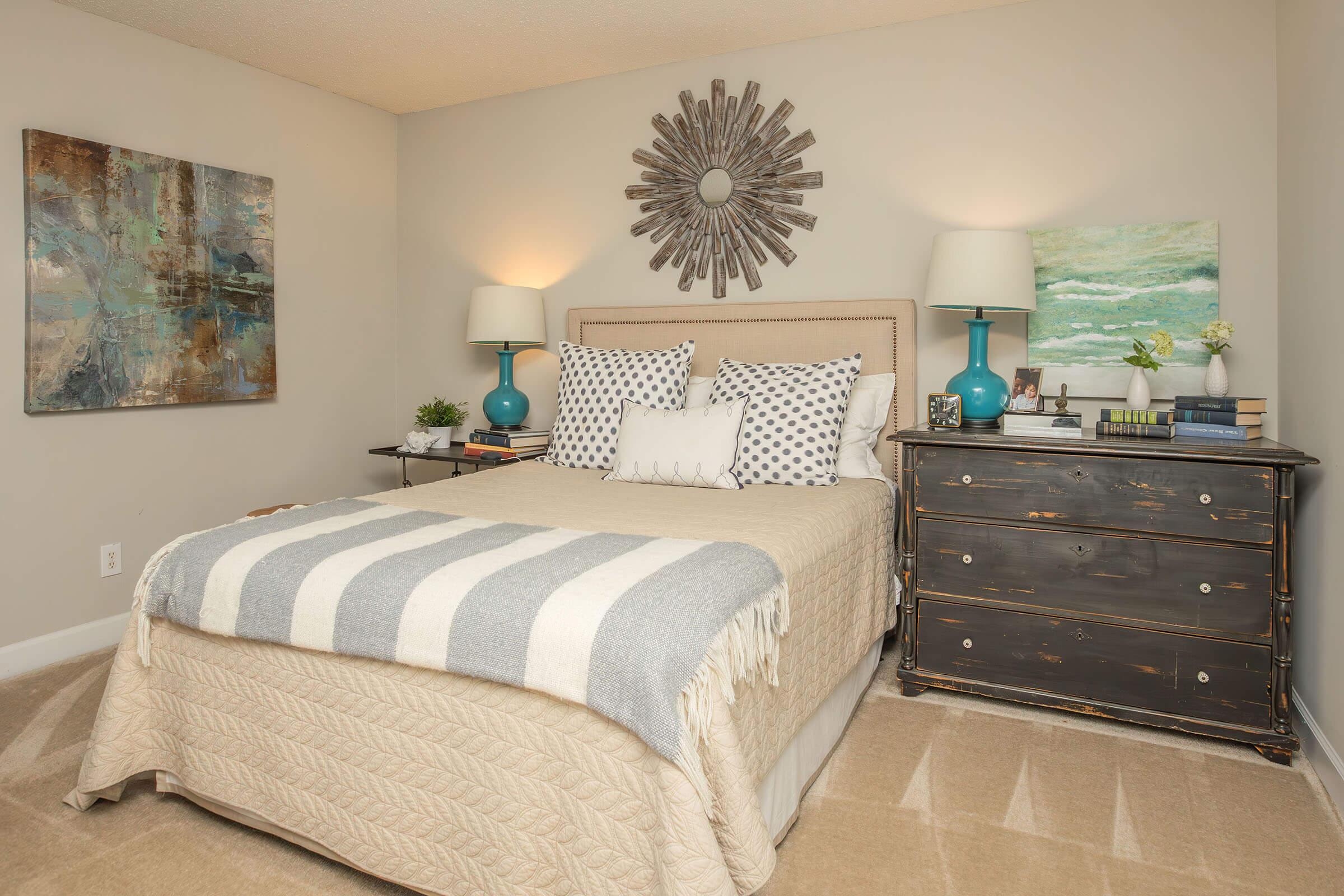 A cozy bedroom featuring a bed with a cream-colored quilt and decorative pillows. There's a round mirror above the headboard, a bedside table with a lamp, and a dark wooden dresser. Artwork hangs on the walls, and the floor has a light carpet, creating a warm and inviting atmosphere.