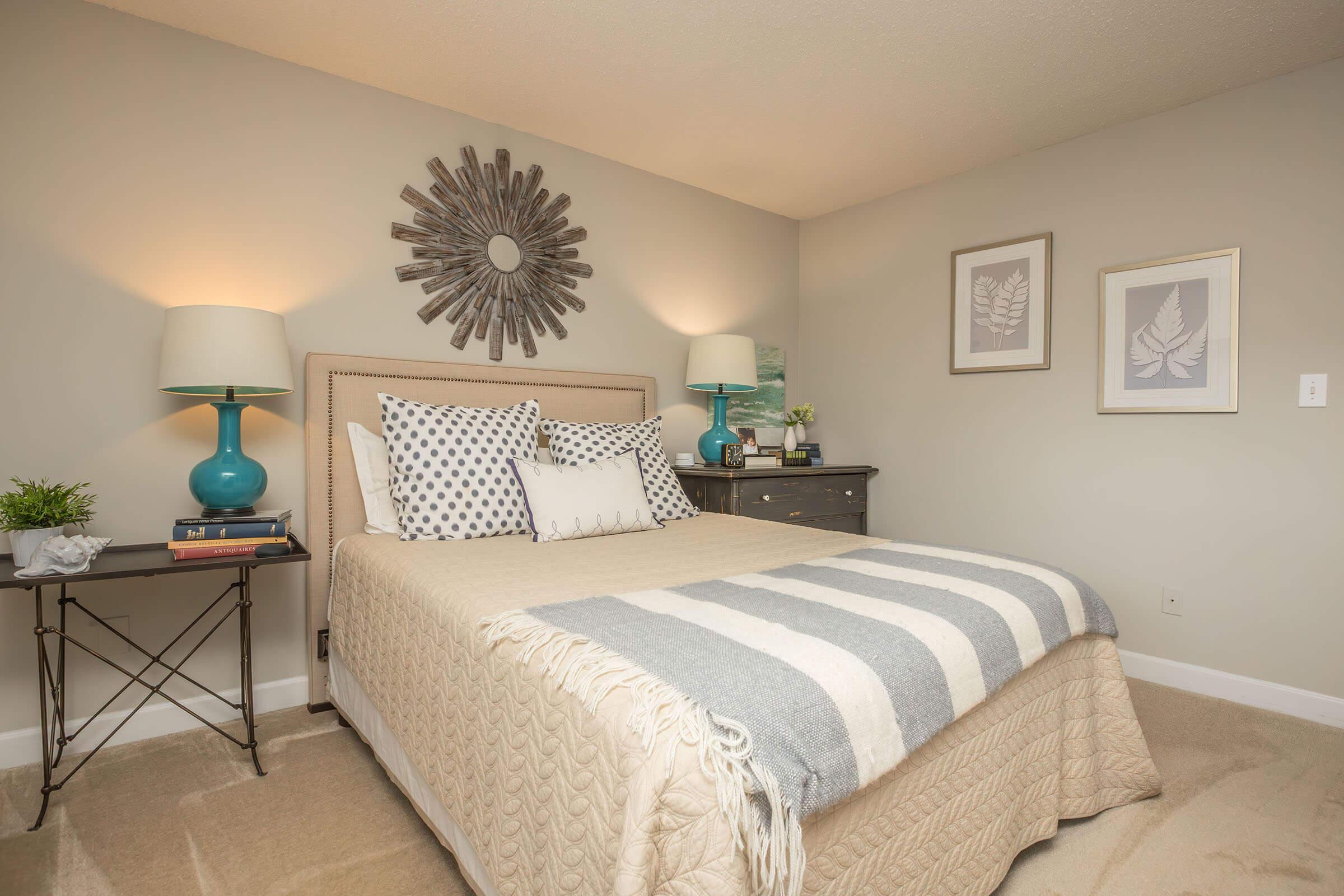 A cozy bedroom featuring a neatly made bed with patterned pillows and a soft throw blanket. Beside the bed, a small table holds a lamp and decorative items. A round mirror and framed artwork adorn the walls, while the neutral color scheme creates a calming atmosphere.