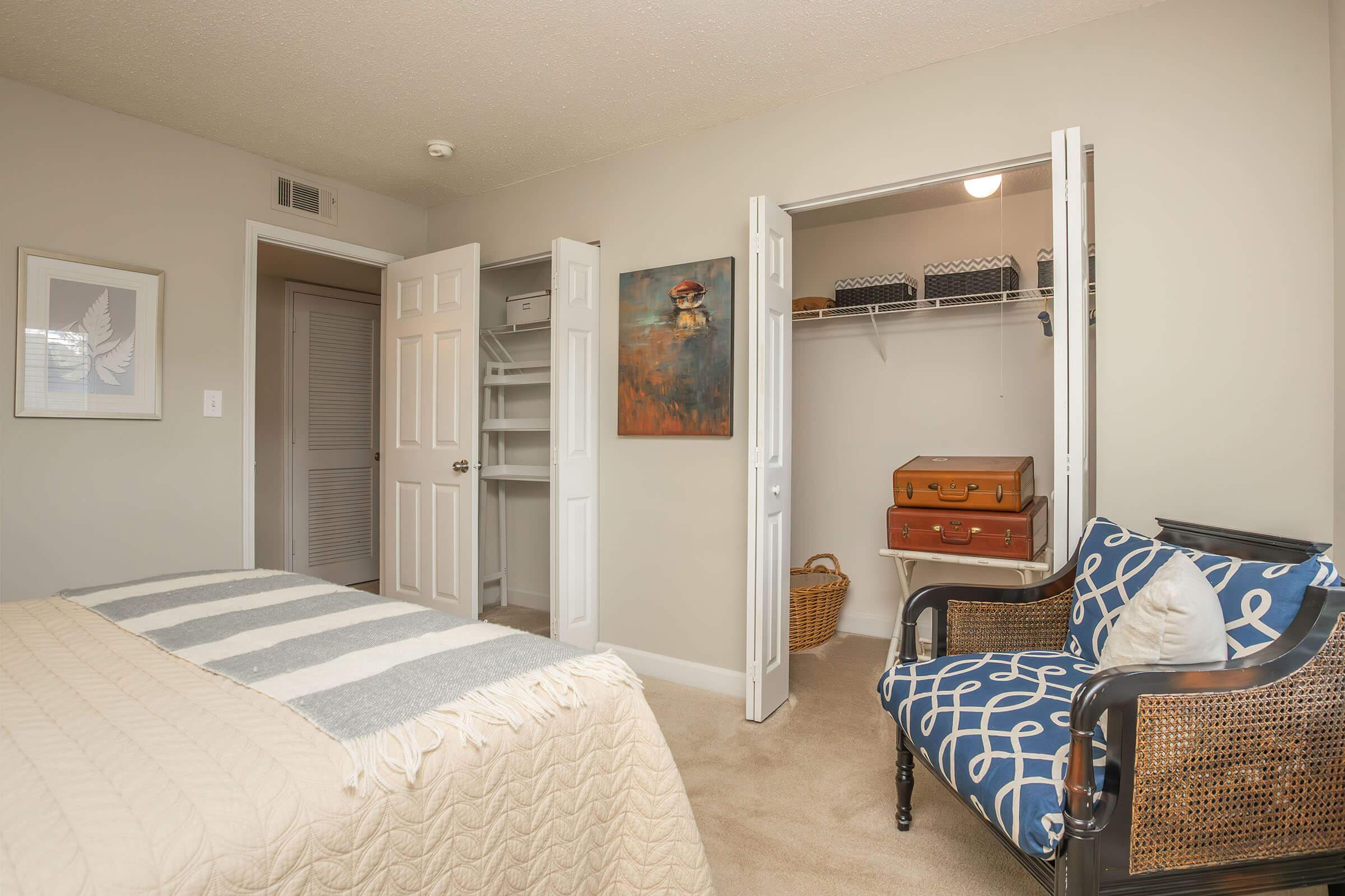 A cozy bedroom featuring a bed with a striped blanket, a decorative chair, and a small closet. The closet has shelves and storing items like suitcases. One wall displays an abstract painting, and the room has neutral-colored walls and carpet, creating a warm and inviting atmosphere.