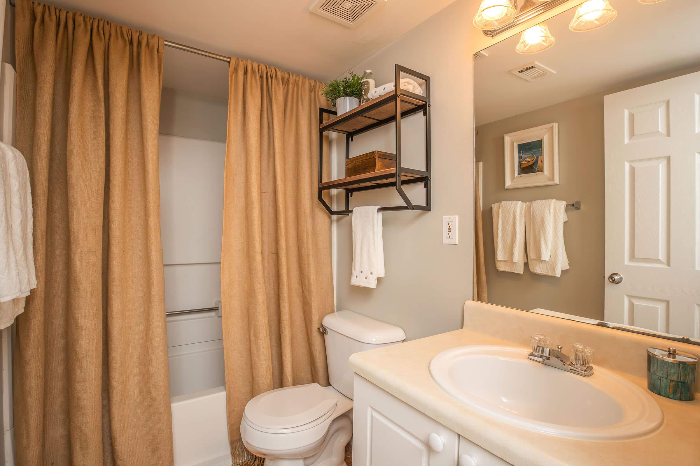 A cozy bathroom featuring a beige shower curtain, a small shelving unit with decorative items, a white toilet, and a sink with a countertop. Soft lighting illuminates the space, creating a warm atmosphere, complemented by neutral wall colors and a framed picture.