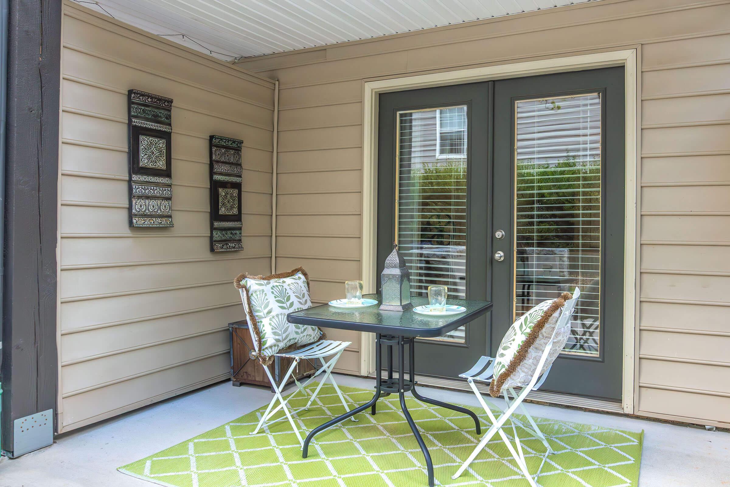 A cozy outdoor patio featuring a small black table with two chairs, set for two with glasses and plates. The walls are light-colored, adorned with decorative panels. There is a green and white patterned rug on the floor, creating a welcoming space for relaxation.