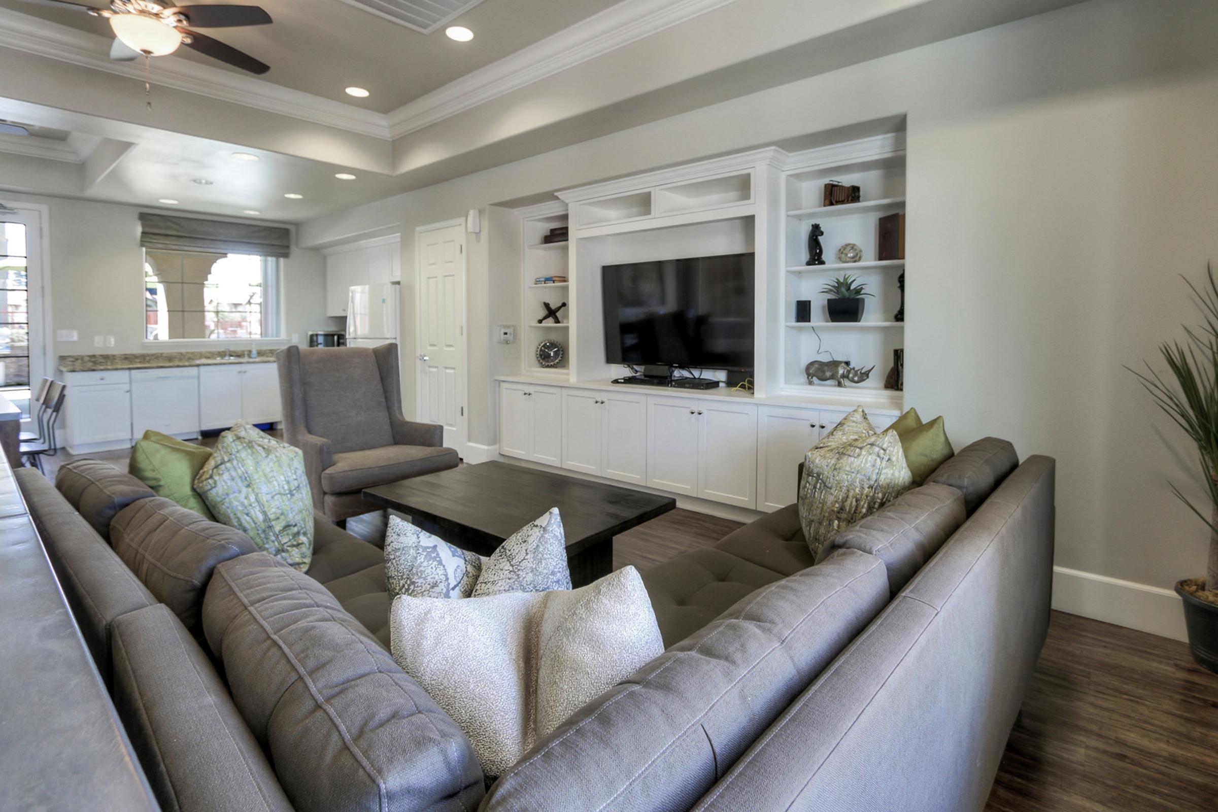 A modern living room featuring a comfortable gray sectional sofa with decorative pillows, a coffee table in the center, and a large flat-screen TV mounted on a white entertainment unit. The space has light walls, wooden flooring, and a ceiling fan, creating a cozy and inviting atmosphere.