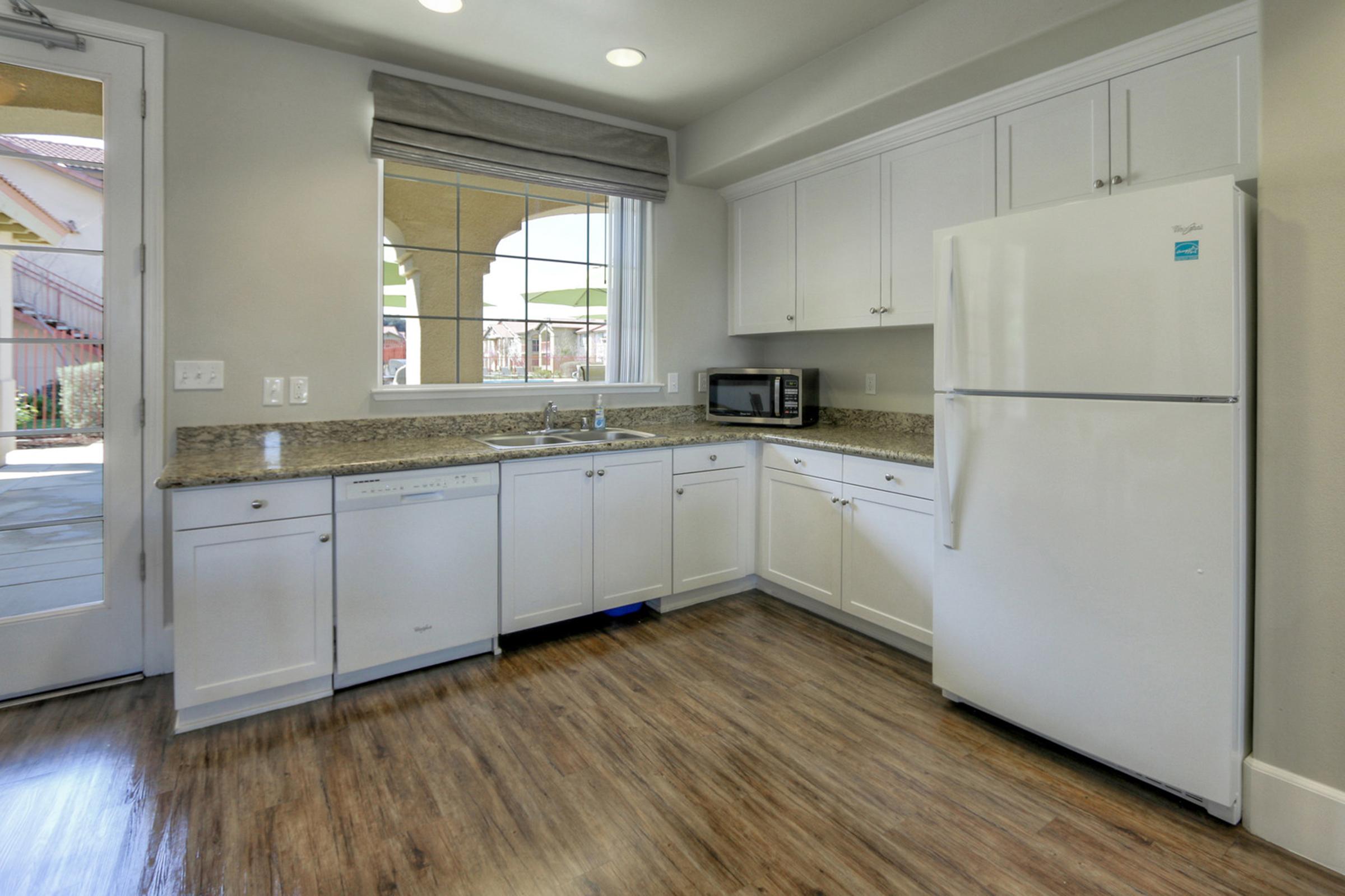Modern kitchen featuring white cabinets, a granite countertop, stainless steel appliances including a refrigerator and microwave, and a large window providing natural light. Wood-look flooring adds warmth to the space, with a view of the outside through the window.