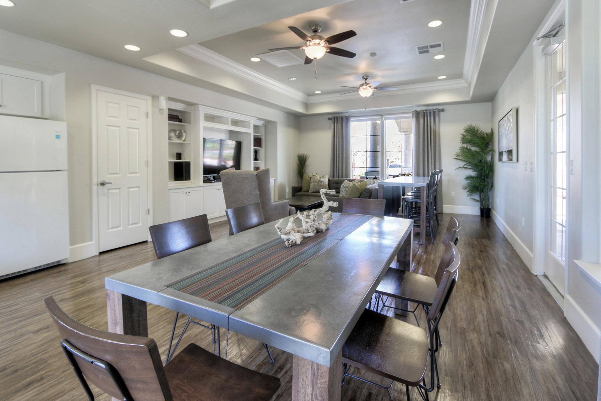 Modern dining area featuring a long metal table with decorative items, surrounded by stylish chairs. The space includes a bright living area with a comfortable sofa, large windows, and a view of greenery. The room's design is open and inviting, with neutral colors and contemporary lighting.