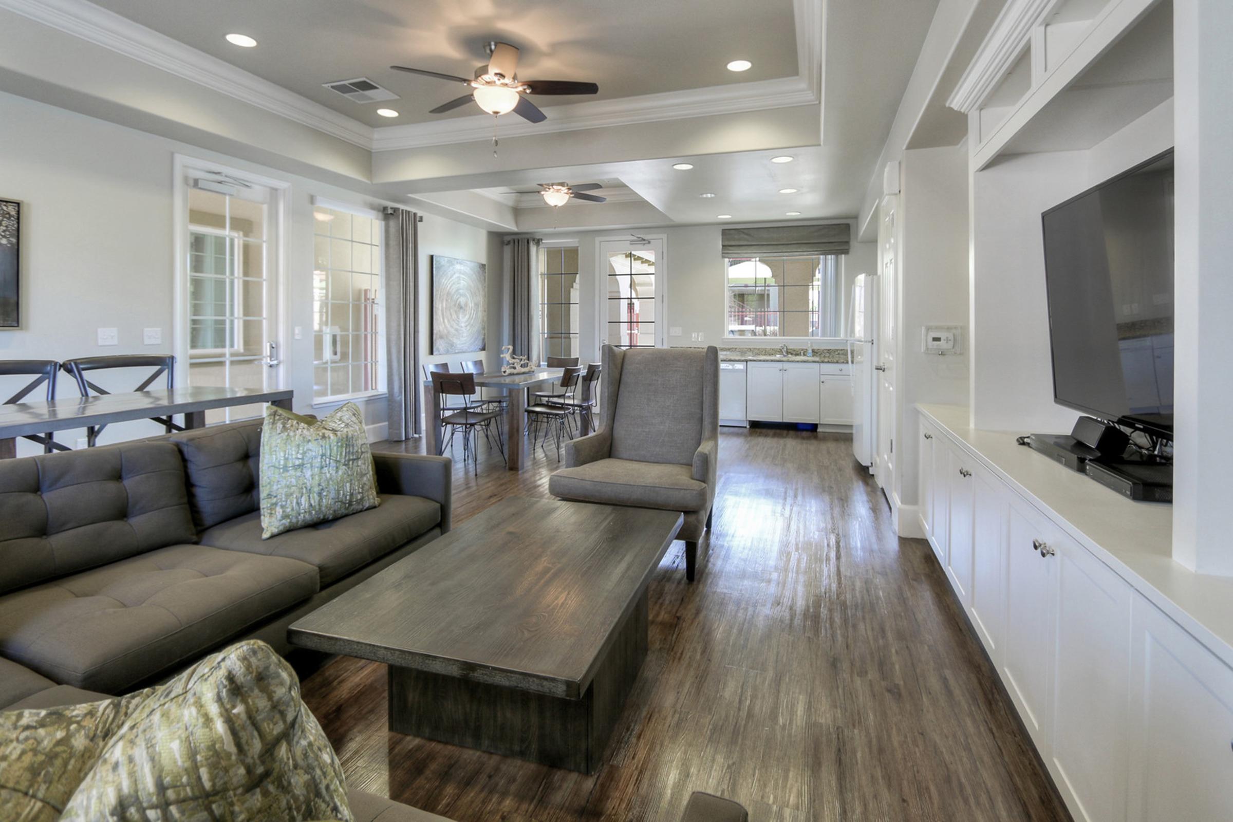 A modern living room featuring a gray sectional sofa with decorative pillows, a wooden coffee table, and a large armchair. The space is well-lit with natural light, showcasing light-colored walls and hardwood flooring. A dining area is visible in the background, along with a kitchen.