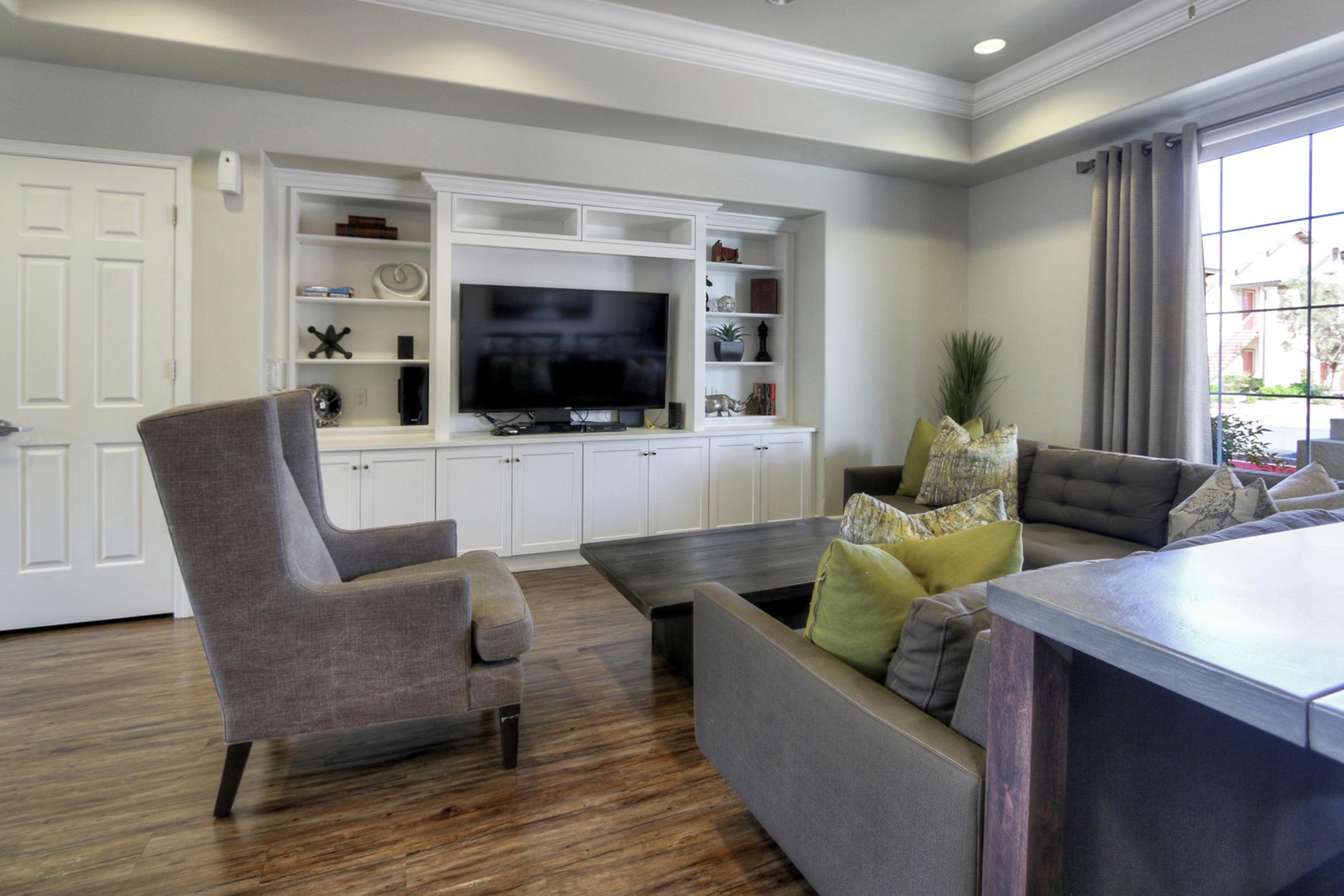 A modern living room with a gray armchair, a sectional sofa with colorful cushions, and a wooden coffee table. There is a large TV mounted on a white entertainment unit, surrounded by decorative shelves. Natural light enters through a large window with gray curtains, and a door is visible on the left.