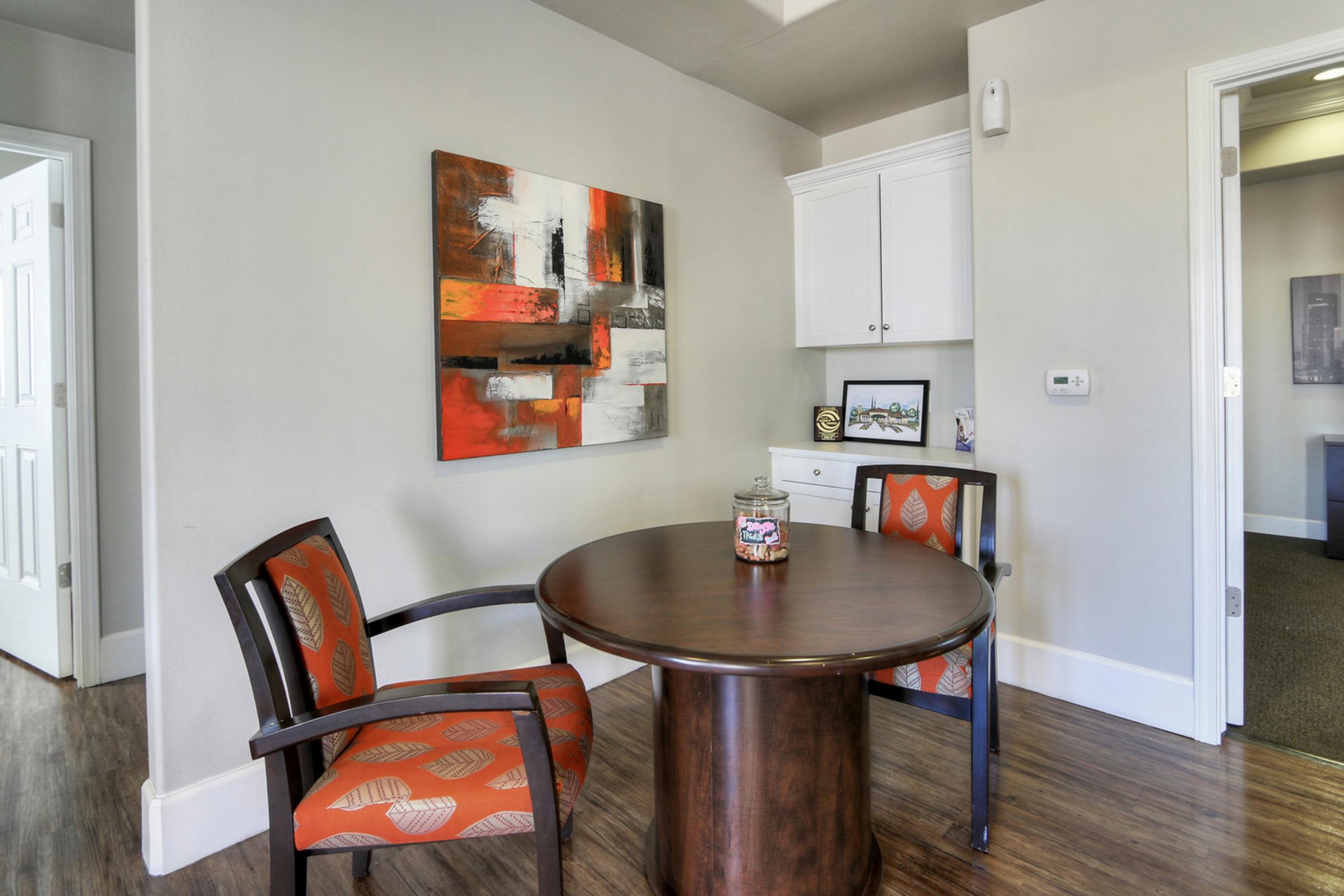 A modern office space featuring a small round table with two chairs. The table has a decorative jar on it, and there is an abstract painting with reds, blacks, and whites on the wall. A white cabinet is visible nearby, along with a door leading to another room. The flooring is wood-like.