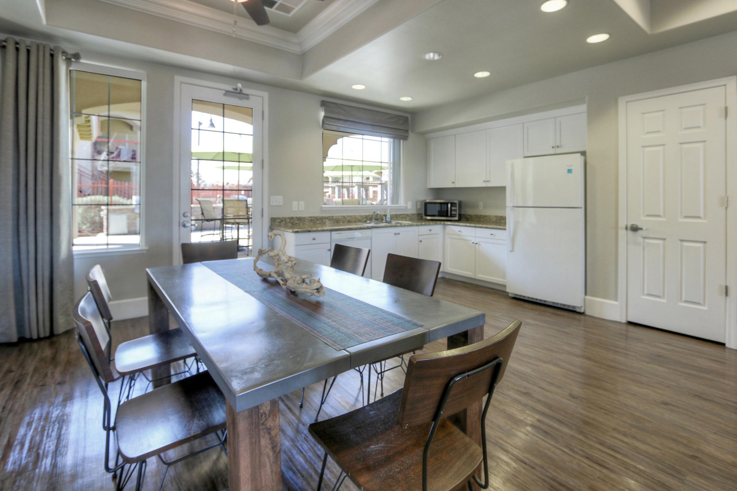 Modern kitchen and dining area featuring a wooden table with metal chairs, a large window letting in natural light, and white cabinetry. There is a white refrigerator and microwave against a wall, with decorative items on the table and grey curtains framing the window.