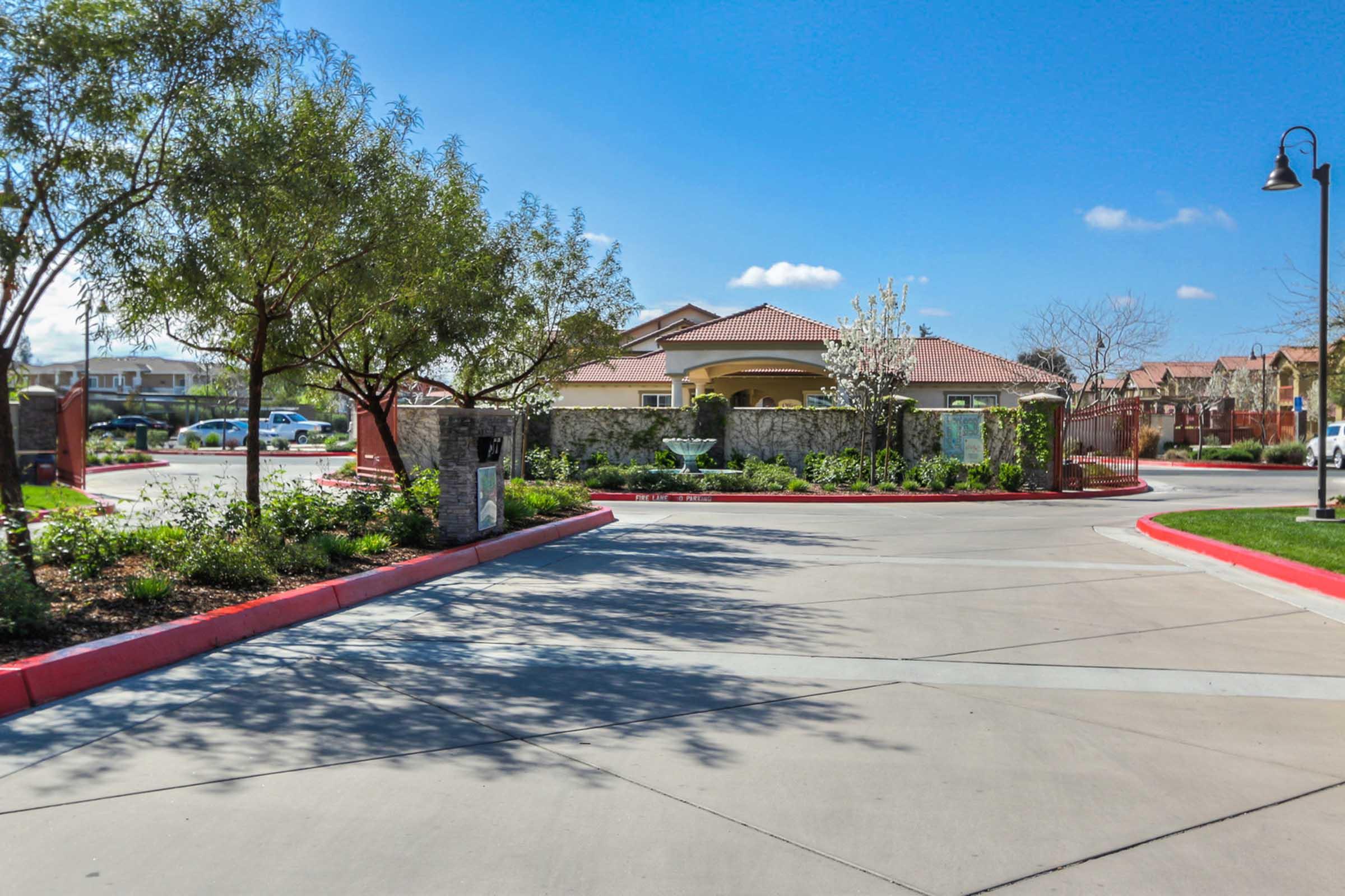 A sunny entrance to a residential community, featuring well-maintained landscaping with trees and flowers, a gated entrance with stone walls, and a clear blue sky. The pathway is paved with a smooth surface leading to a building in the background, with vehicles parked along the side.