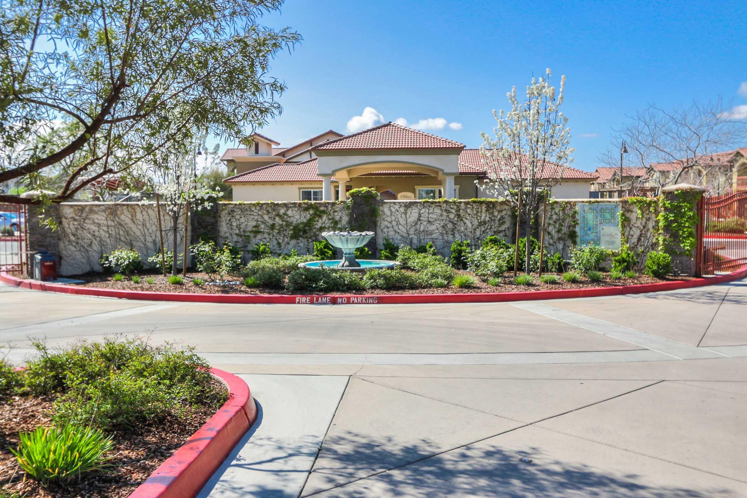 A decorative fountain surrounded by lush greenery and blooming flowers at the entrance of a residential complex. The building in the background features a red-tiled roof and is partially obscured by a stone wall. Clear blue skies with a few clouds complete the scenic view.