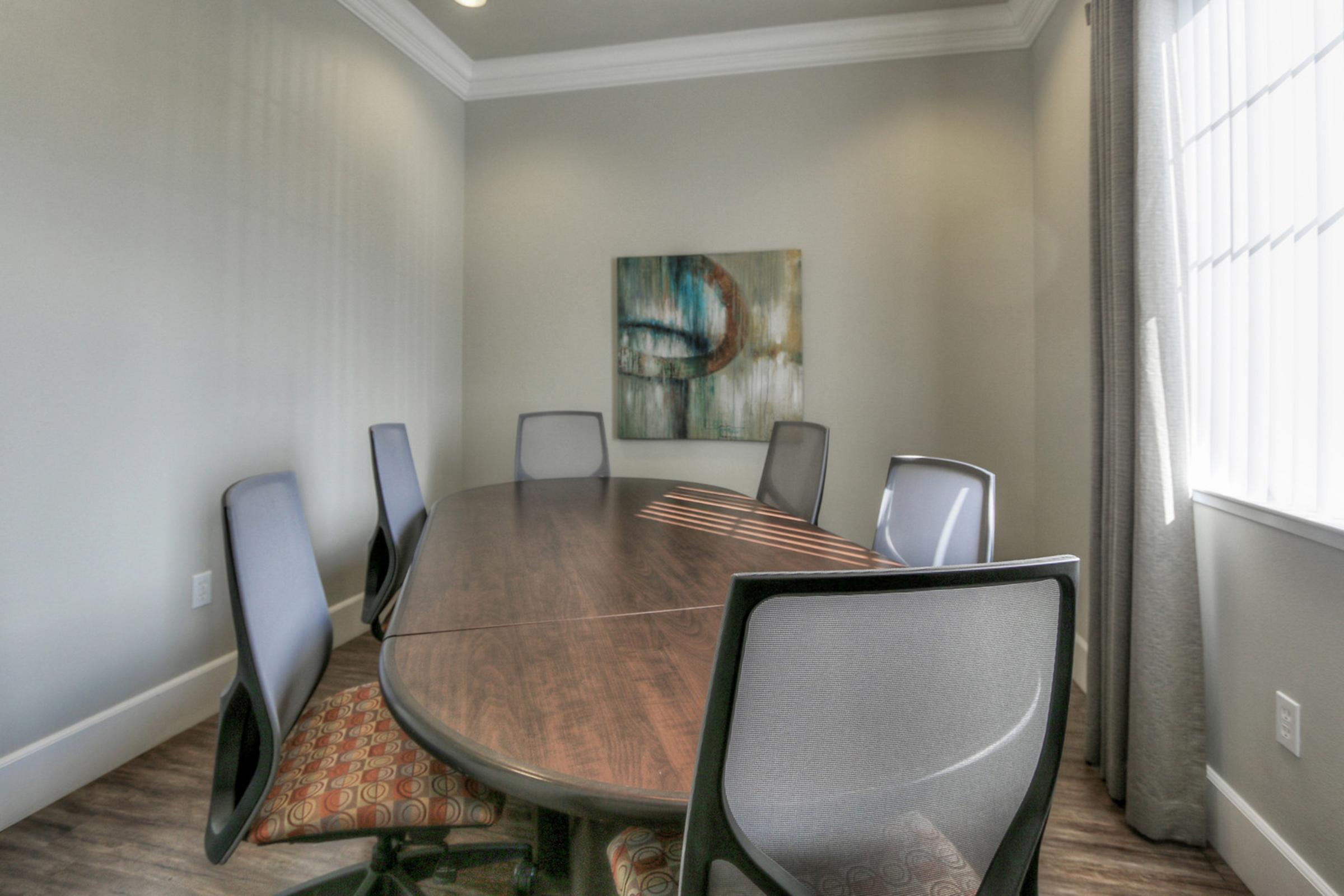 A modern conference room featuring a large oval wooden table surrounded by ergonomic chairs. The walls are painted in neutral tones, and there's a piece of abstract art hanging on the wall. Natural light filters through a large window covered with sheer curtains.