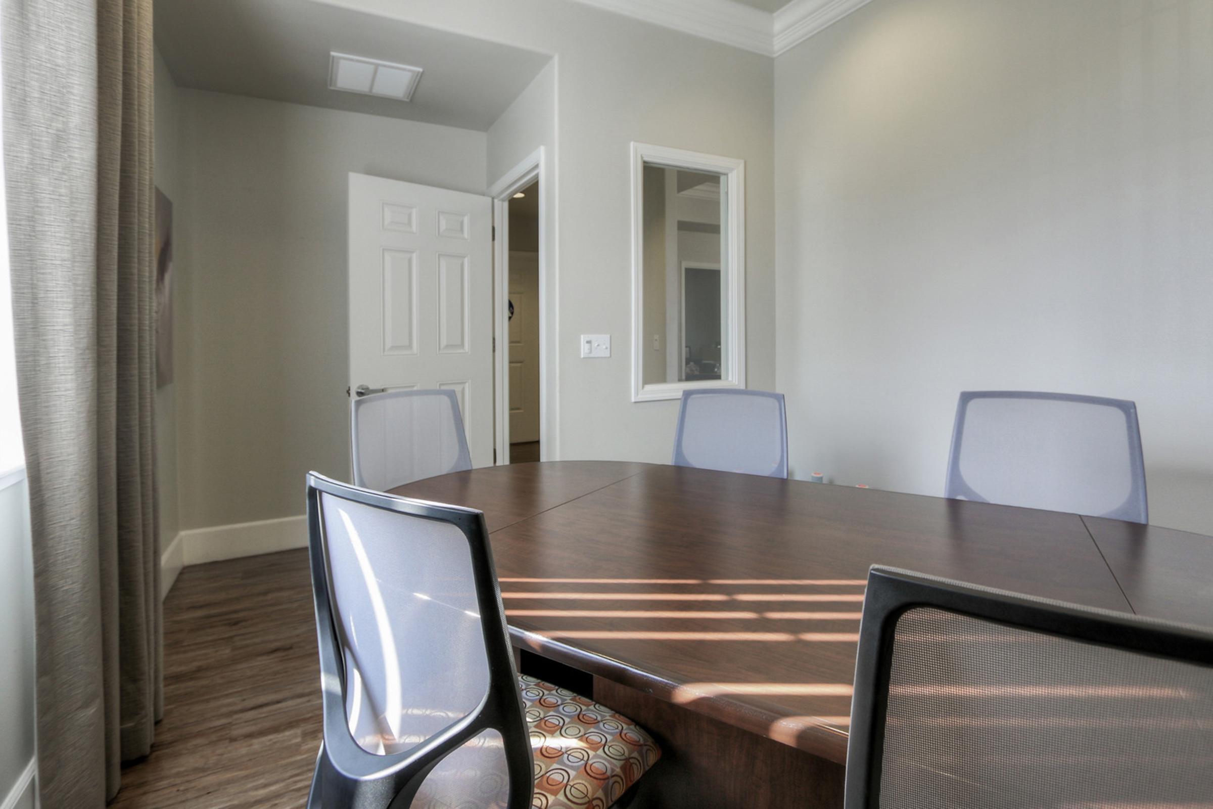 A modern conference room featuring a large oval wooden table surrounded by four stylish chairs. Soft sunlight streams through a window, casting shadows on the floor. The room has neutral-colored walls and a door leading to another area, with a large mirror visible in the background.