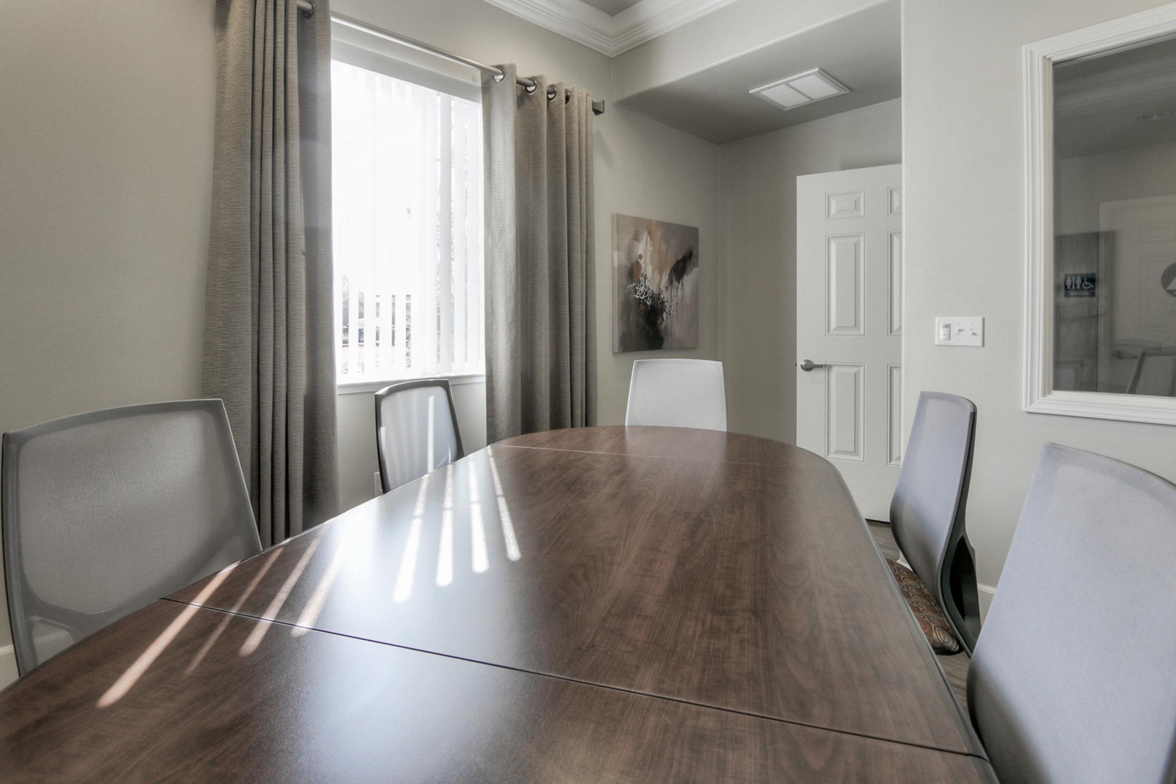 A modern conference room featuring a long wooden table surrounded by four gray ergonomic chairs. Natural light filters in through a window with sheer curtains, casting lines on the table. A closed door and a framed artwork are visible in the background, creating a professional and inviting atmosphere.