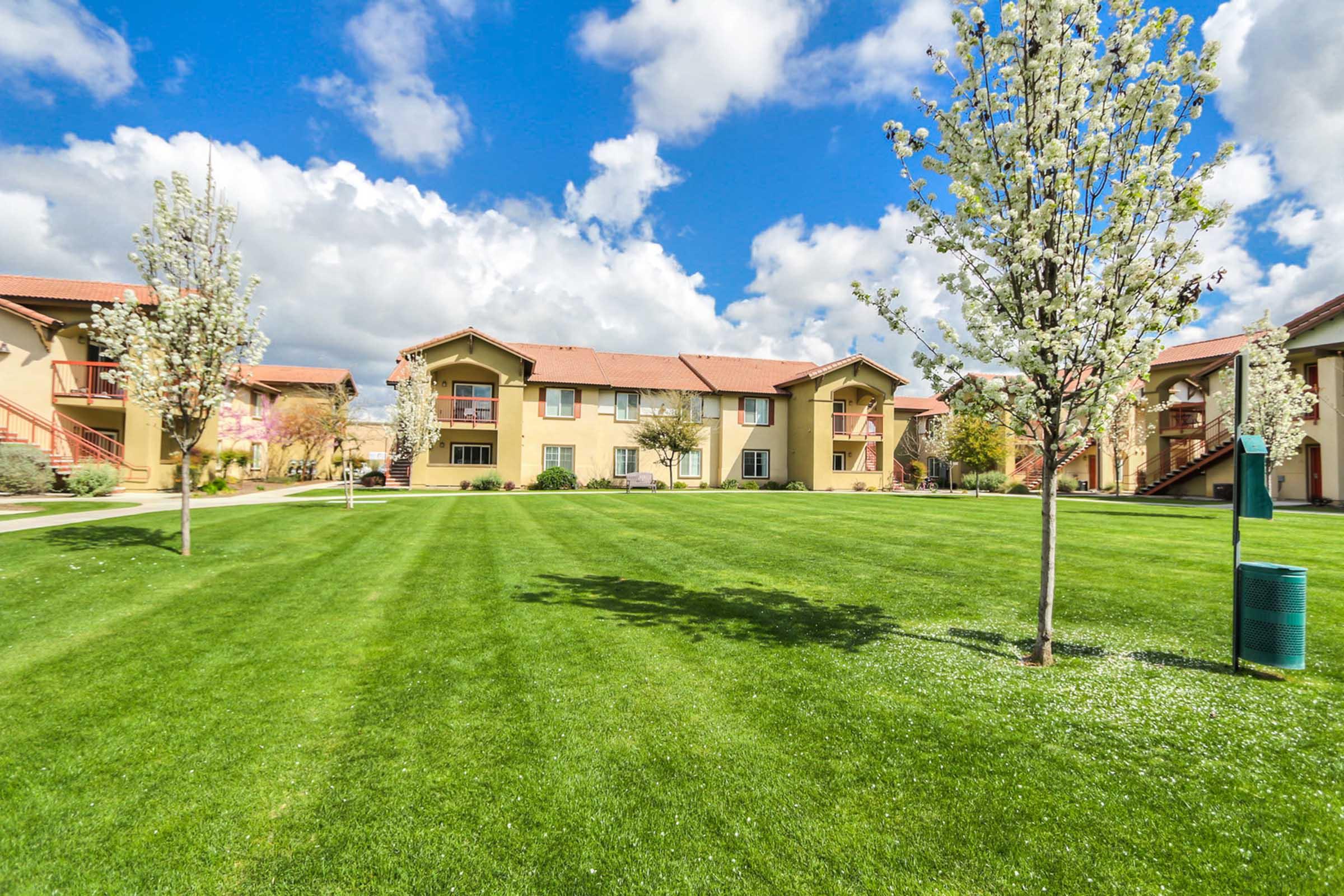 A well-maintained residential complex featuring two-story buildings with a mix of pastel colors. The foreground has a lush green lawn with small trees, while the background displays a vibrant blue sky dotted with fluffy clouds. The setting is peaceful and inviting, ideal for outdoor activities or relaxation.