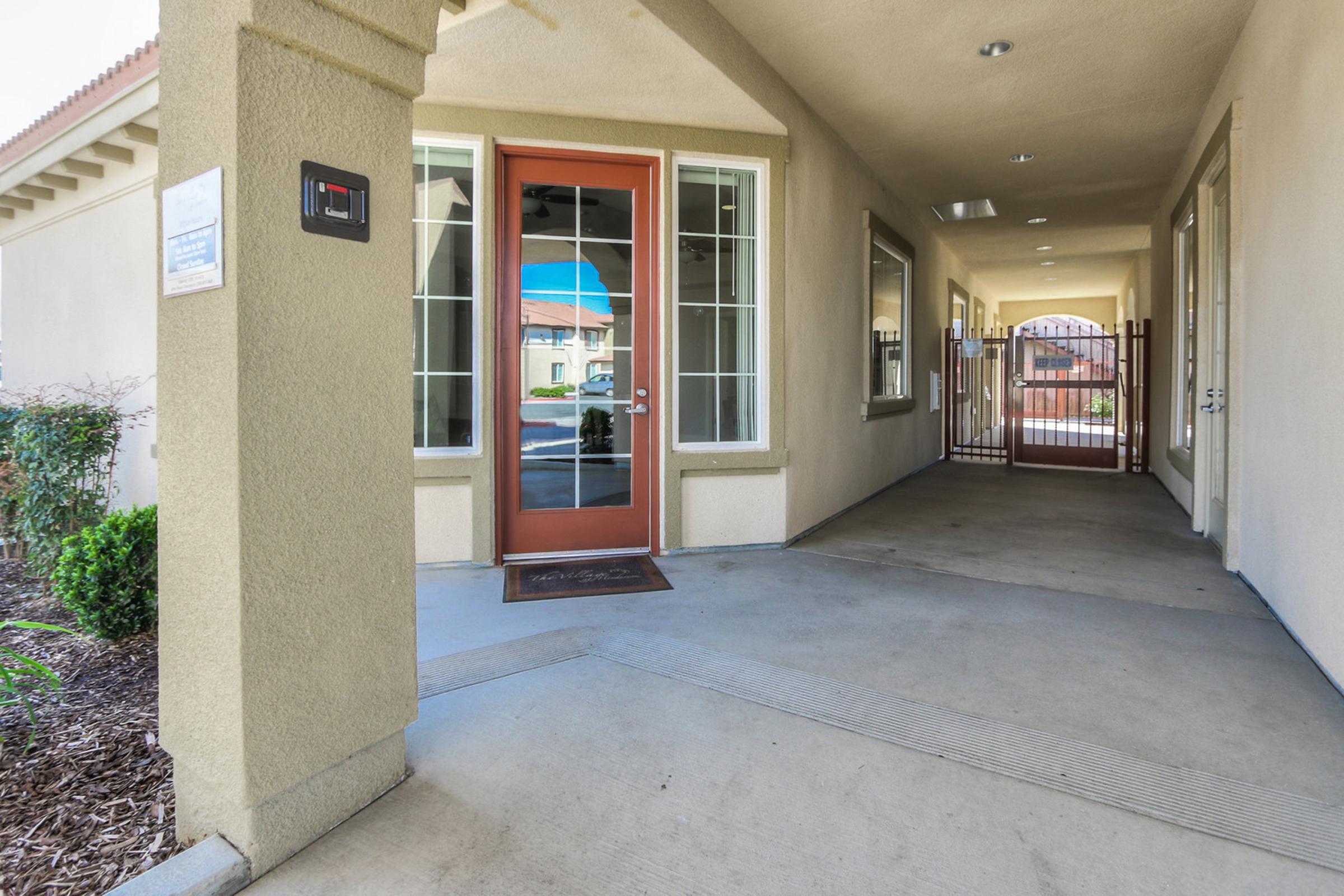 A covered entryway featuring a glass door with a reddish-brown frame. The pathway is lined with small shrubs, and a gate is visible on the right side. Natural light floods the space, highlighting the clean and tidy surroundings. The area is well-maintained and offers access to a community or building.