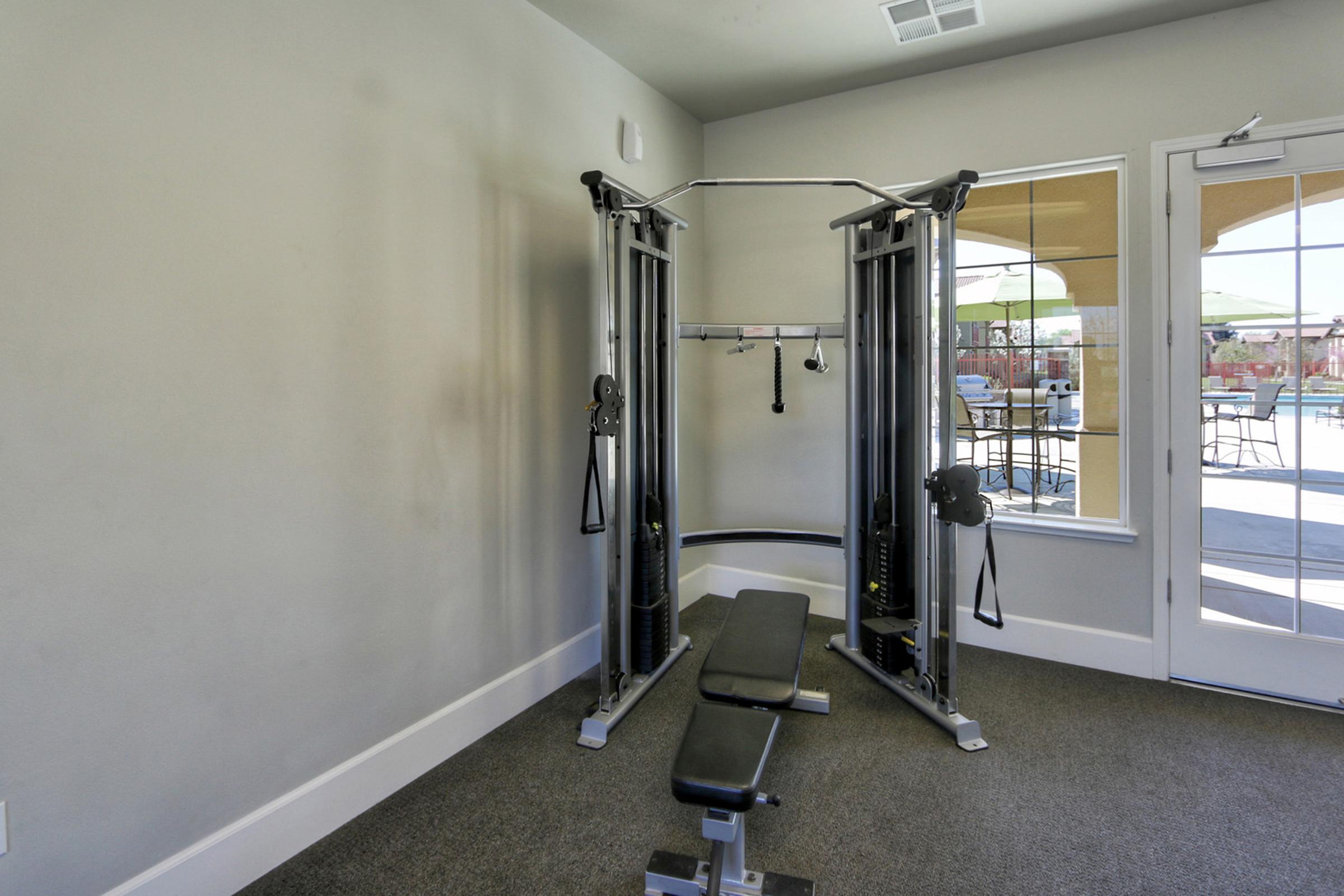 A fitness room features a cable machine with a bench in front of it. Large windows allow natural light, providing a view of an outdoor area with patio seating. The walls are neutral-colored, and the flooring is carpeted.