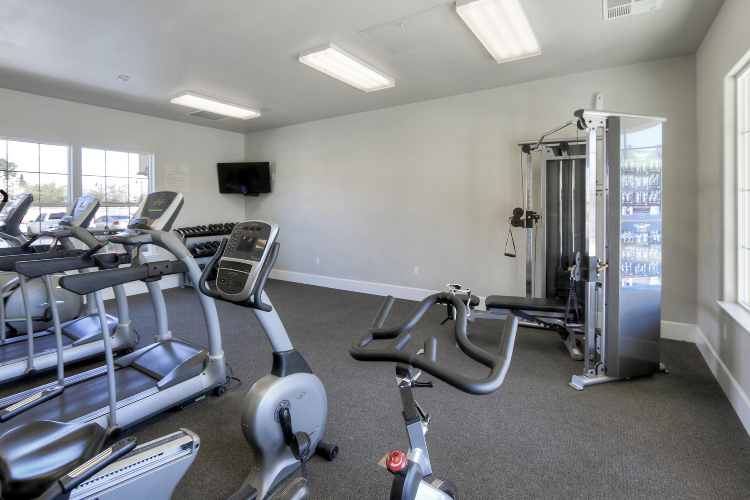 A bright, modern gym interior featuring several exercise machines, including treadmills and stationary bikes. On one side, there's a multi-station weight machine, and on the opposite wall, a TV is mounted. The walls are painted in a light color, contributing to the spacious feel of the room.