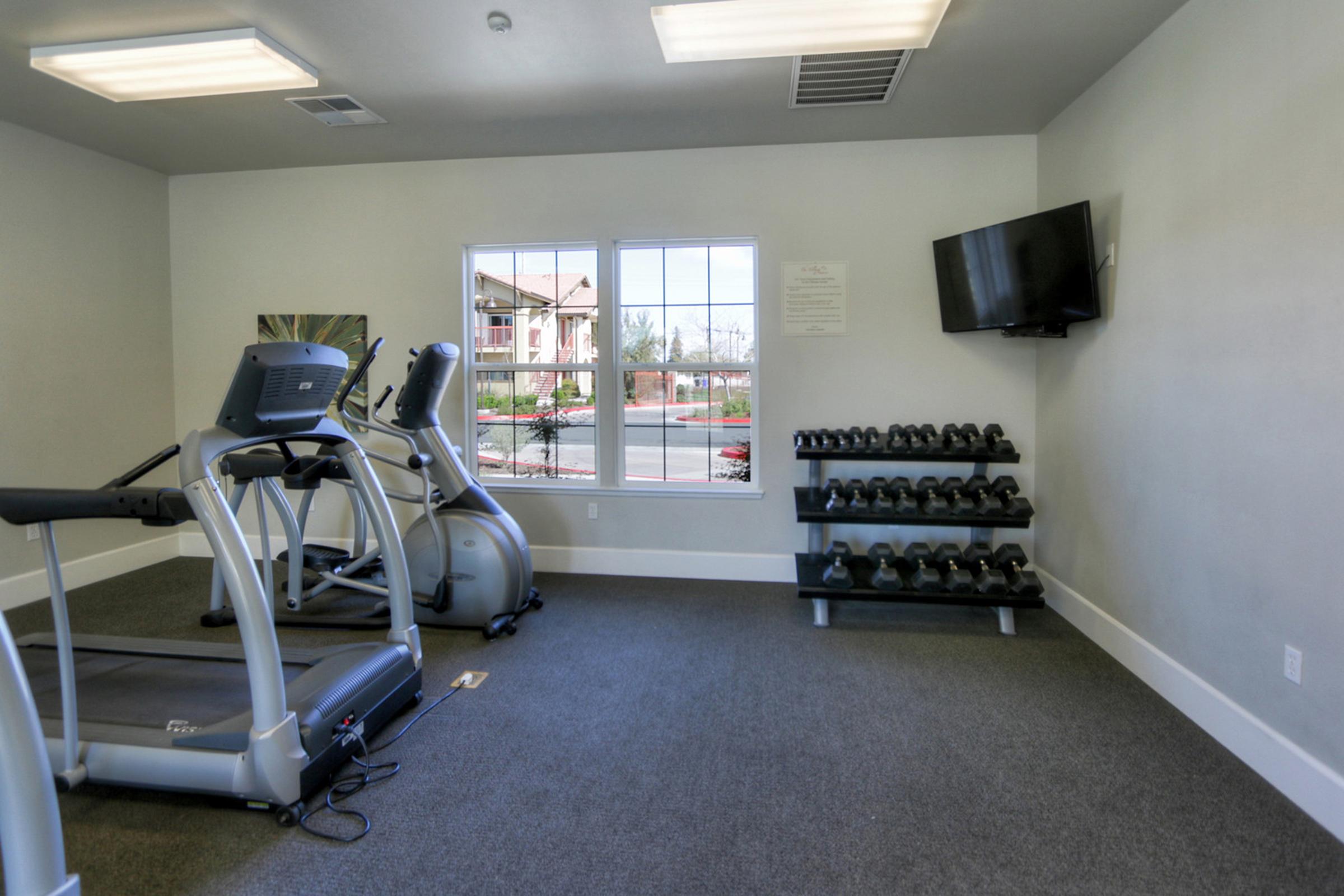 A small exercise room featuring two cardio machines, a wall-mounted TV, and a rack of black dumbbells. The space has light-colored walls and is well-lit with natural light coming in from a window. Fitness equipment is neatly arranged on a carpeted floor.