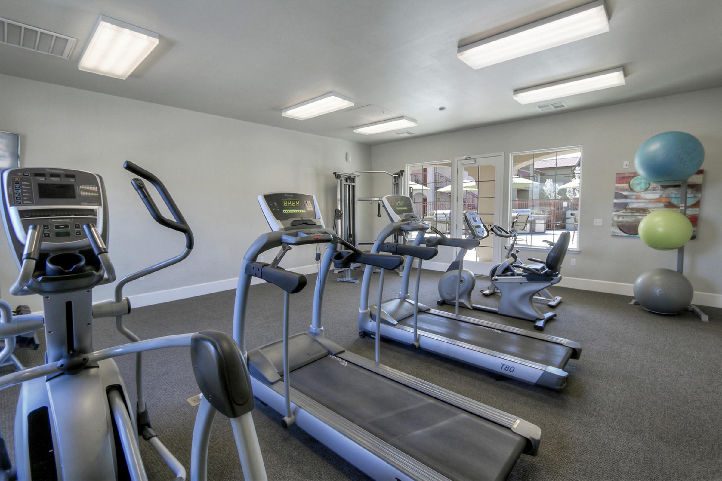 A modern gym interior featuring various fitness equipment, including treadmills and ellipticals. The space is well-lit with overhead lights and large windows, and there are exercise balls in the corner. The flooring is carpeted, and the environment appears clean and inviting for workouts.