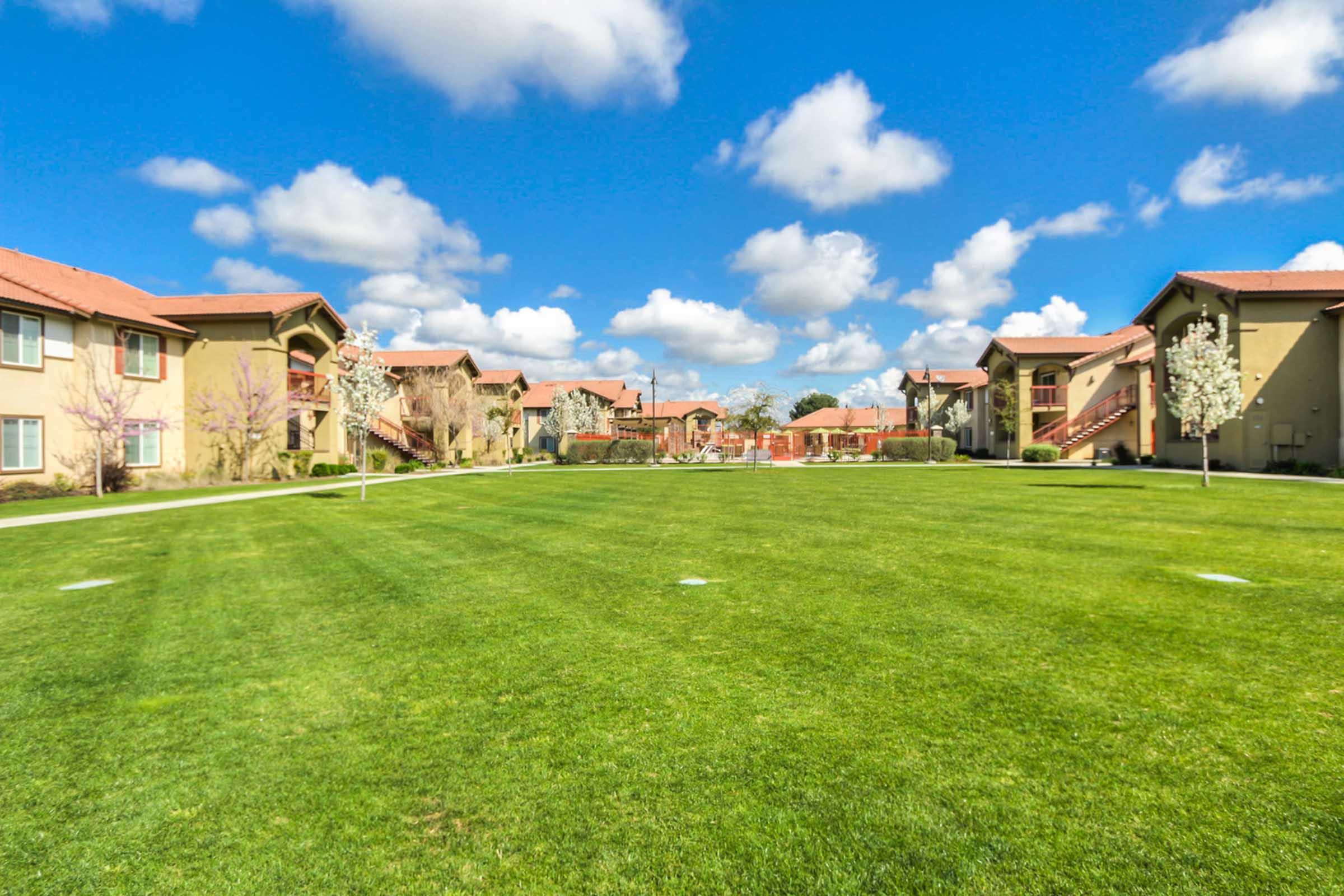 A wide view of a landscaped apartment complex featuring green grass, trees, and sunny skies with fluffy white clouds. The image shows several buildings with balconies in a well-maintained setting, creating a peaceful residential atmosphere.