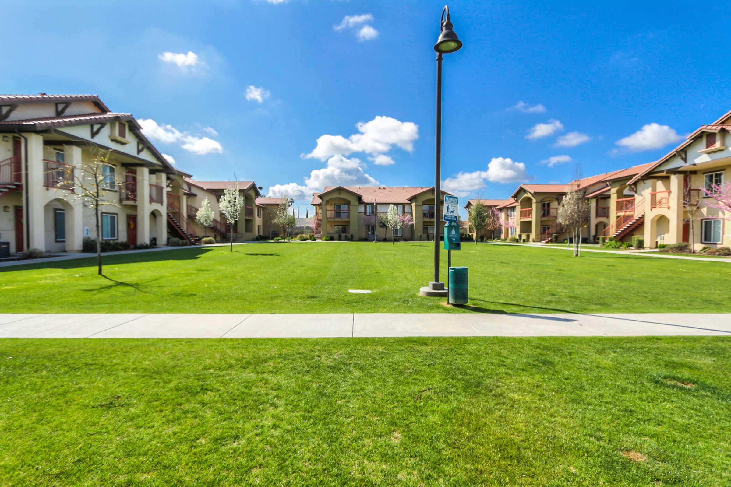 A well-maintained grassy area with several residential buildings in the background under a clear blue sky with fluffy clouds. The scene includes a pathway for walking and a lamppost, creating a serene atmosphere in a residential community.