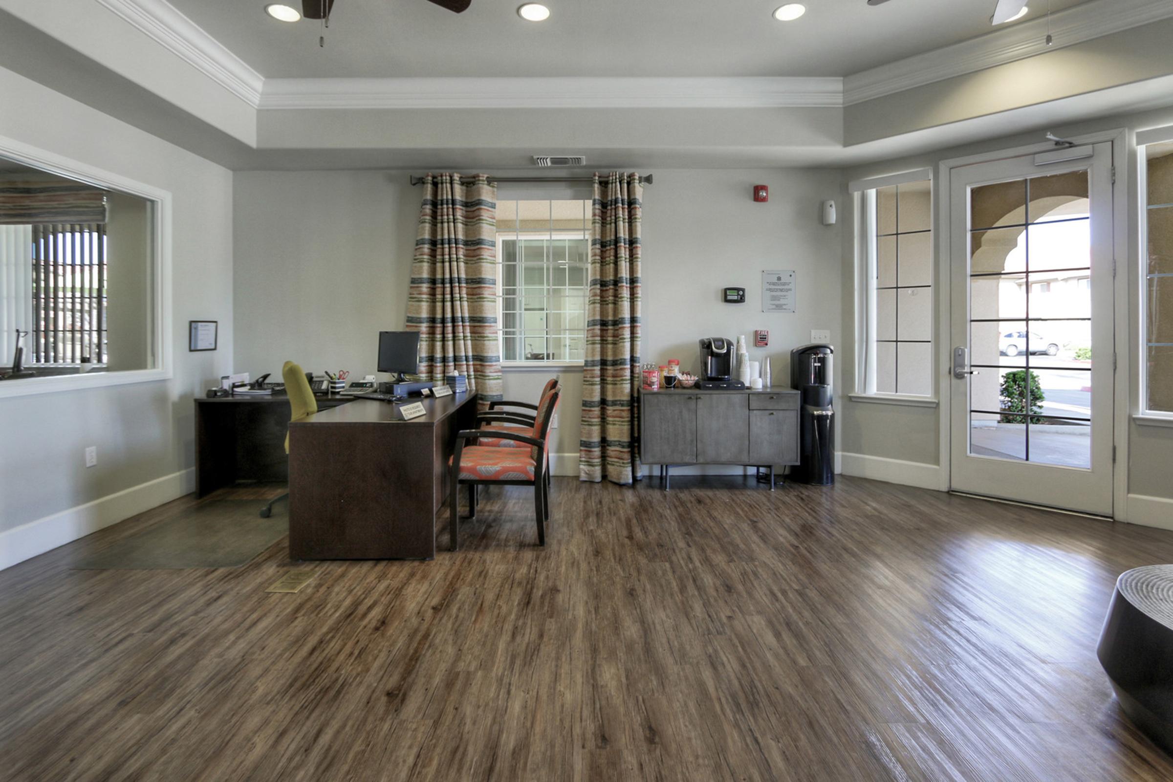 Interior of an office space featuring a reception area with two desks, chairs, and a coffee machine. The walls are painted light colors, and there are large windows with decorative curtains. The floor is a wooden laminate, creating a welcoming atmosphere.