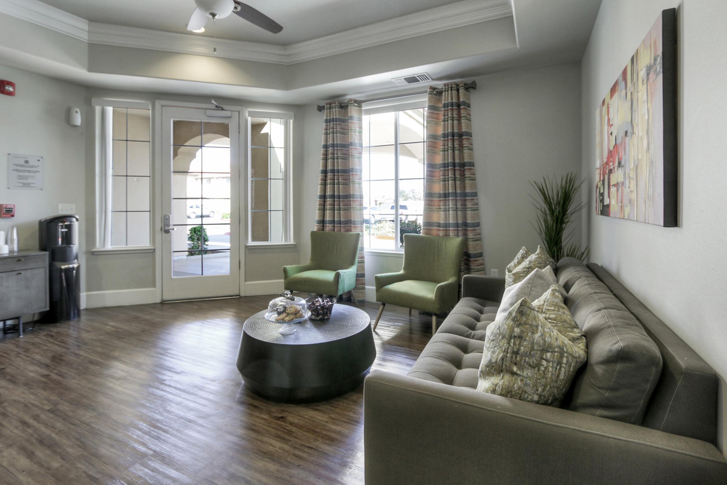 A modern living room featuring a gray sofa with patterned pillows, two green chairs, a round coffee table with decorative items, and large windows with sheer curtains. The space is well-lit and designed for comfort, with a potted plant and artwork on the wall, creating a welcoming atmosphere.