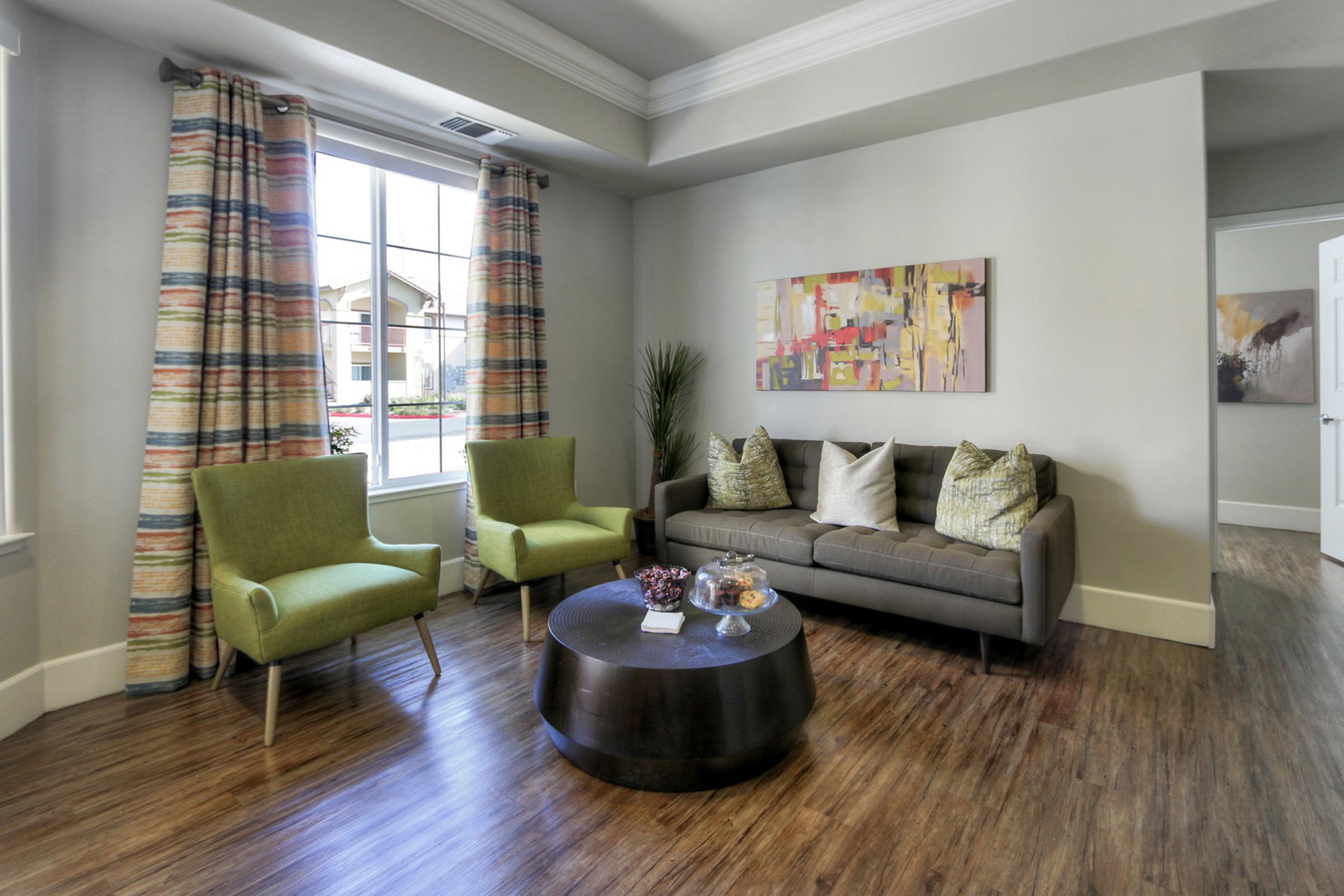 A modern living room featuring a gray sofa with decorative pillows, two green armchairs, large windows with colorful curtains, and a round coffee table with a bowl of snacks. The walls are painted in soft tones, and artwork is displayed on one wall, adding a vibrant touch to the space.