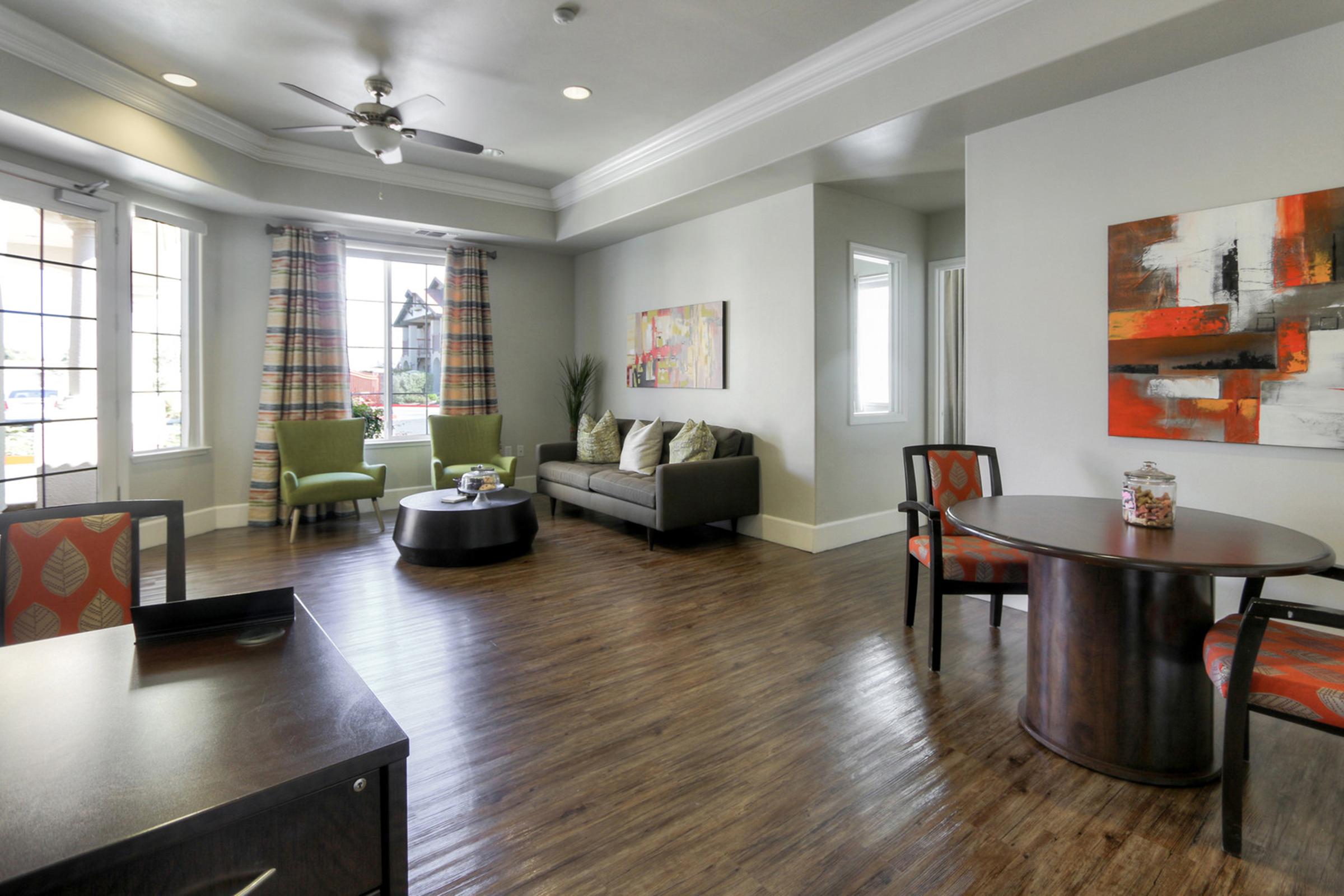A modern, well-lit living area featuring a gray sofa with decorative pillows, two green armchairs, a round coffee table, and a dark dining table with chairs. Large windows with plaid curtains allow natural light, and a colorful abstract painting hangs on the wall. The floor is made of dark wood.