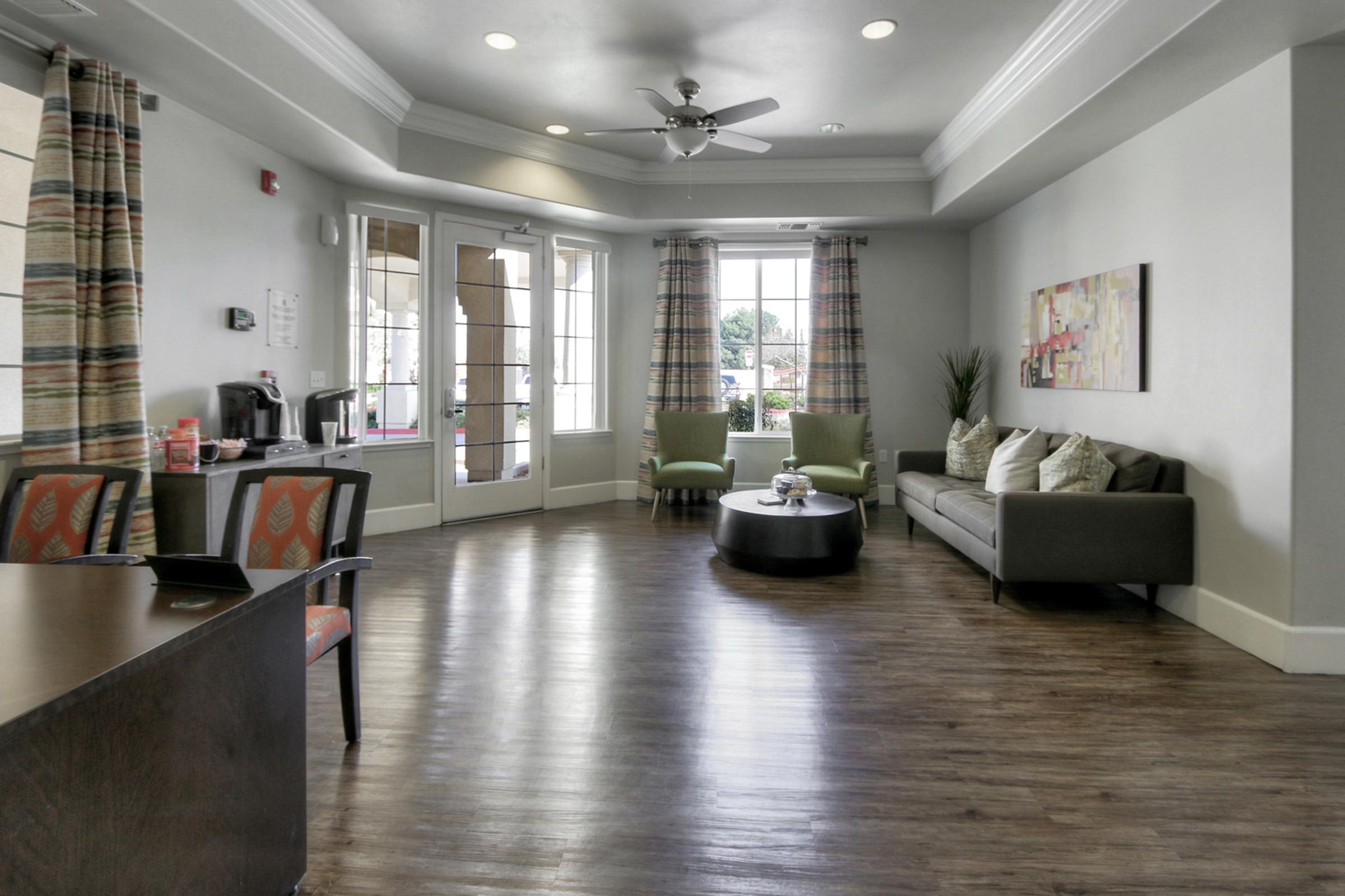 A modern lounge area with large windows allowing natural light. Features include a gray sofa with colorful pillows, a round coffee table, and armchairs. There’s a wall-mounted artwork, a fan on the ceiling, and a refreshment station. The flooring is wooden and the walls are painted in light colors, creating a welcoming atmosphere.